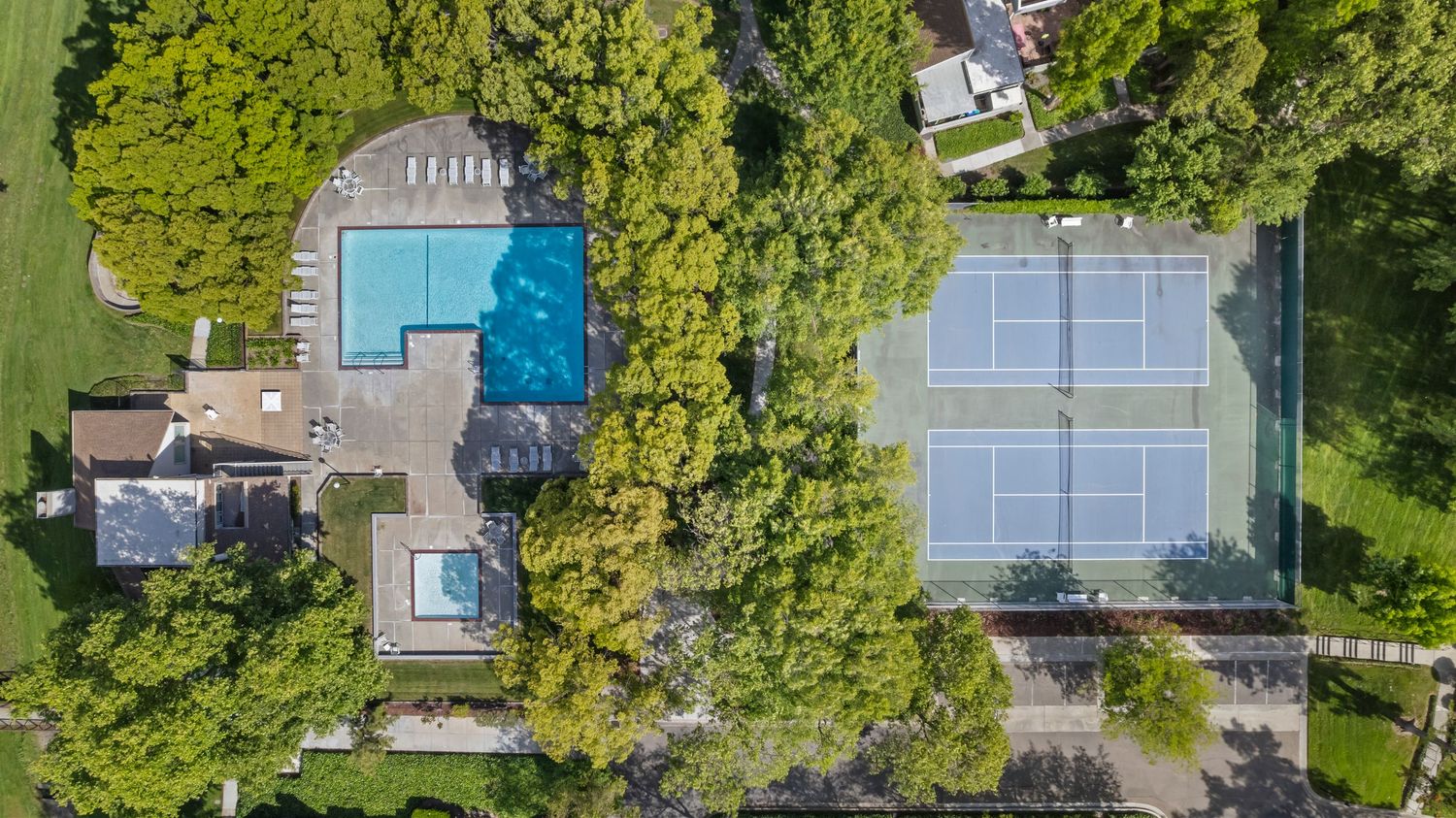 Aerial view of community amenities featuring a sparkling swimming pool and blue tennis courts surrounded by mature trees.
