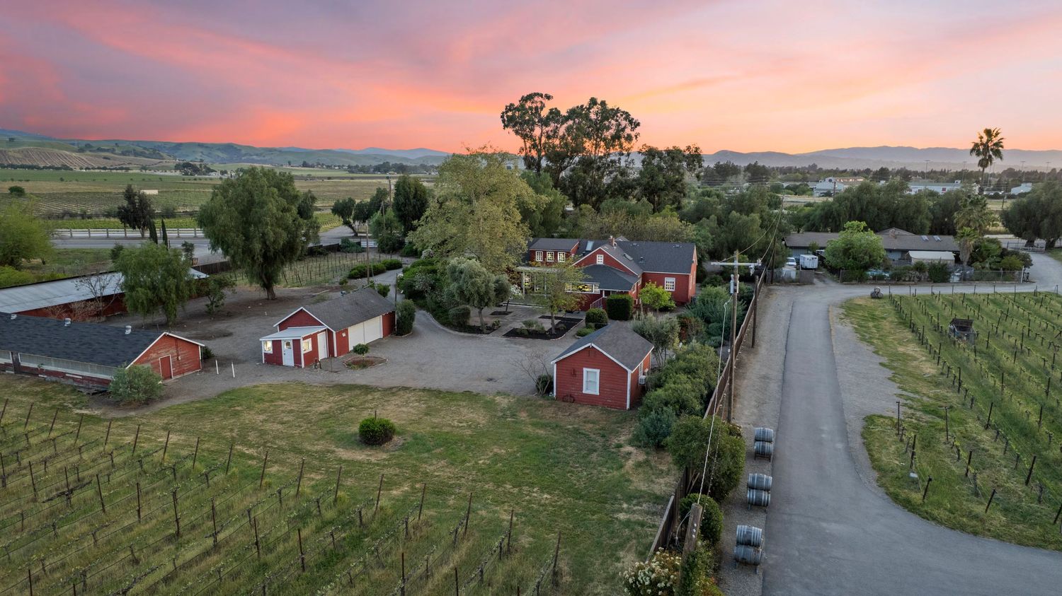 Aerial view of rural vineyard property with red barn buildings, mature trees, and valley farmland at sunset.