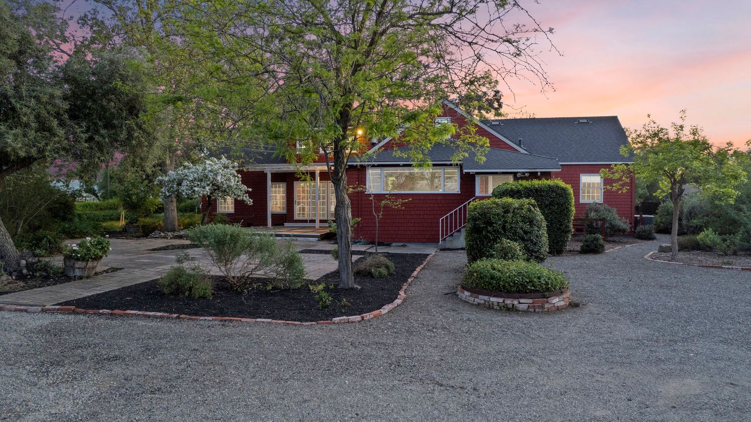 Charming red brick ranch home at dusk with gravel driveway, mature trees, and manicured garden beds.