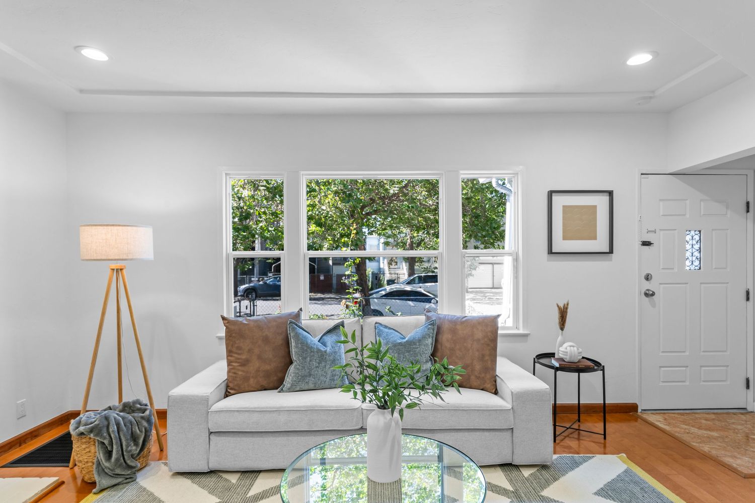 Bright living room with tray ceiling, crown molding, white sofa, hardwood floors, and large window with garden view.
