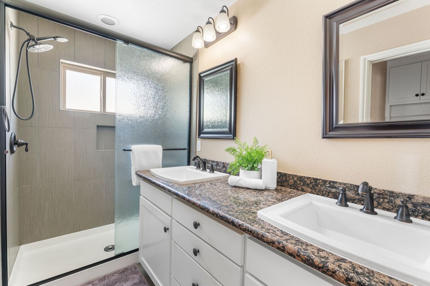 Modern bathroom with double sinks, dark granite countertop, white cabinets, and glass-enclosed shower.