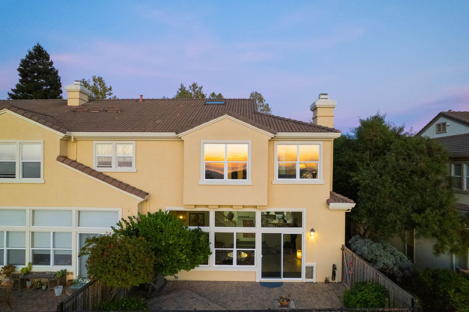Stunning two-story stucco home with large windows reflecting golden sunset light, featuring a tiled roof and landscaped patio.