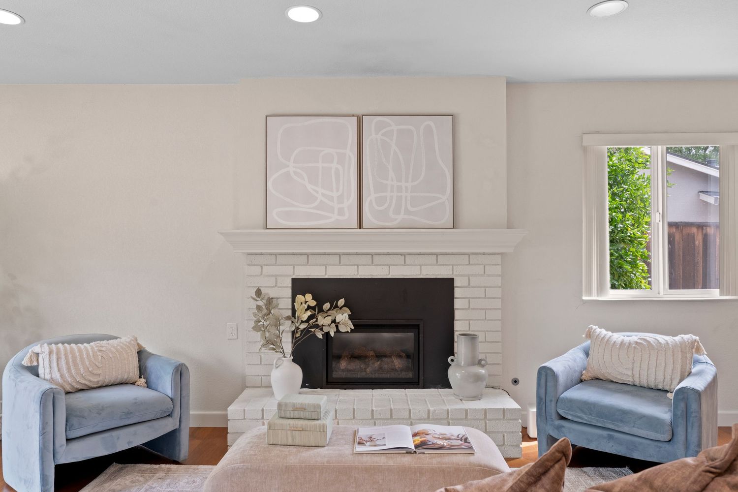 Bright living room with white brick fireplace, two blue velvet chairs, and abstract art above the mantel.