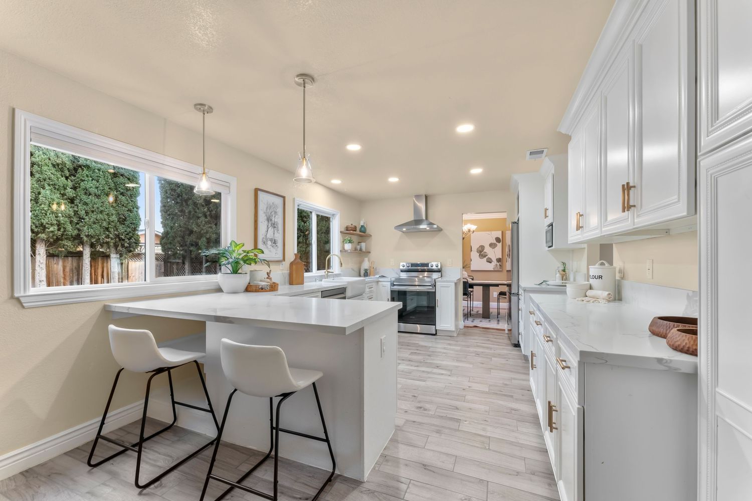 Modern white kitchen with island seating, pendant lights, quartz countertops, stainless range, and light wood-look flooring.