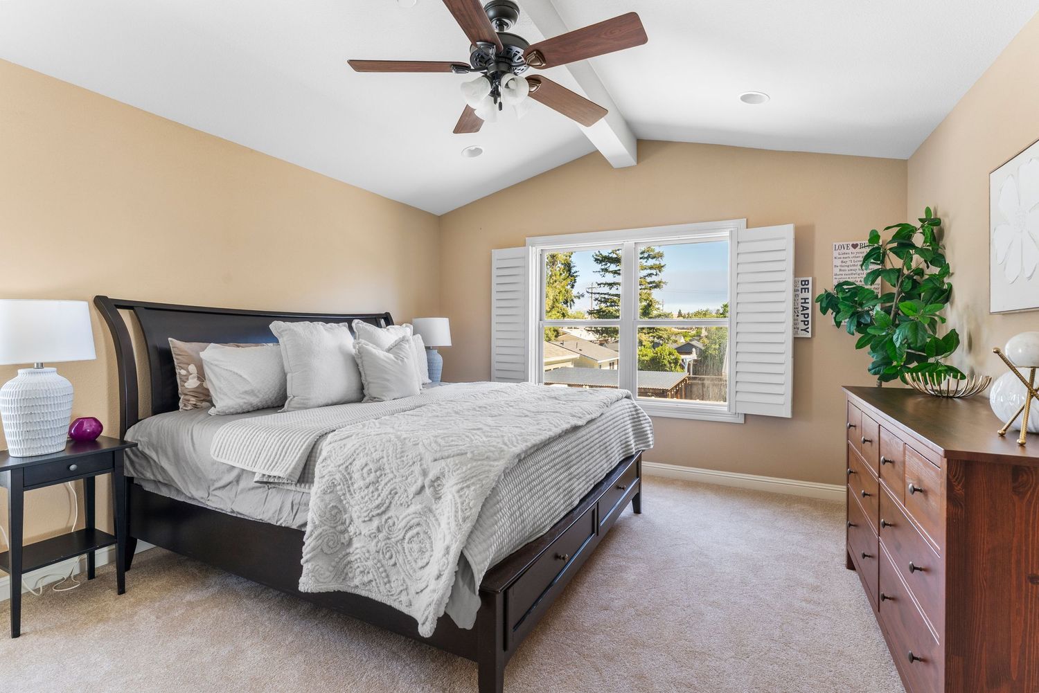 Spacious master bedroom with vaulted ceiling, ceiling fan, large window, gray bedding, and wood dresser.