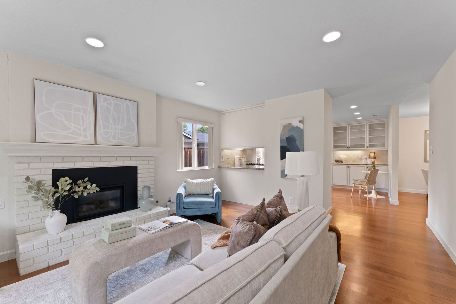 Bright living room with brick fireplace, neutral sofa, hardwood floors, and open view to kitchen with white built-in shelving.