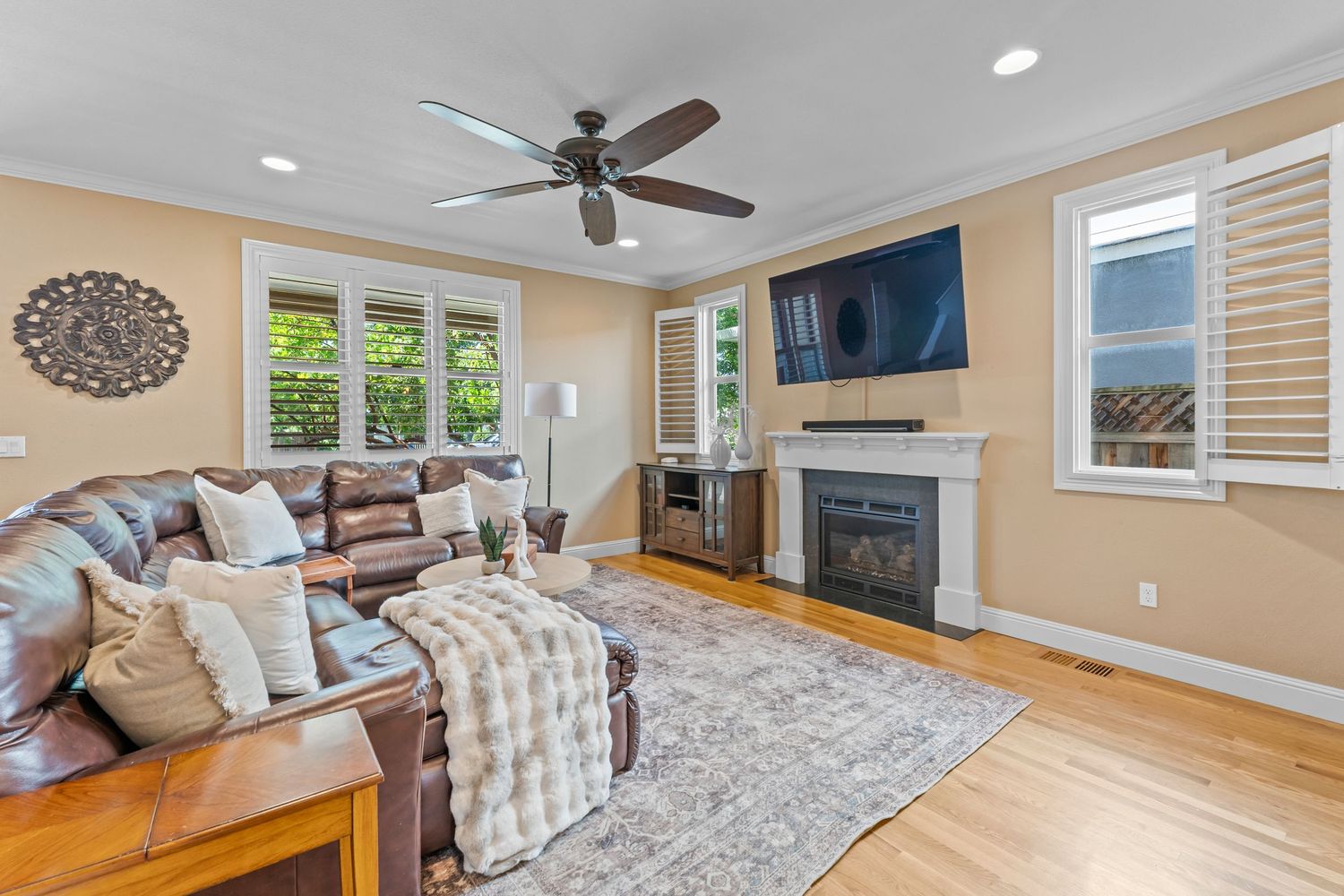 Cozy living room with brown leather sofa, hardwood floors, ceiling fan, fireplace, and large windows.