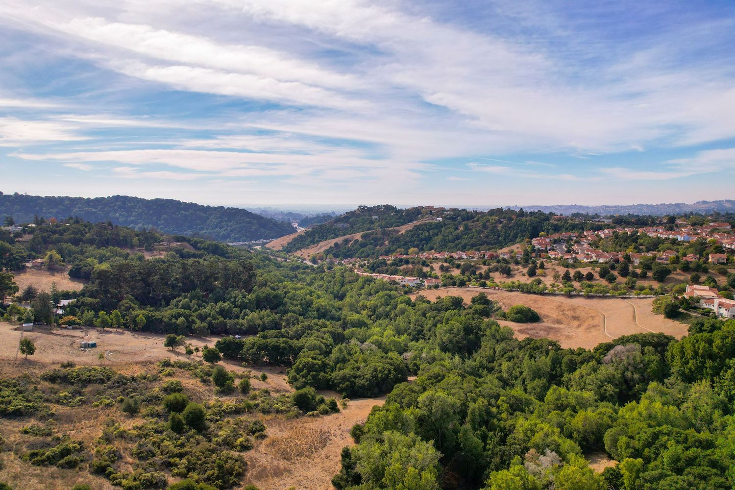 Panoramic aerial view of rolling hills, green valleys, and suburban neighborhoods under a partly cloudy blue sky.