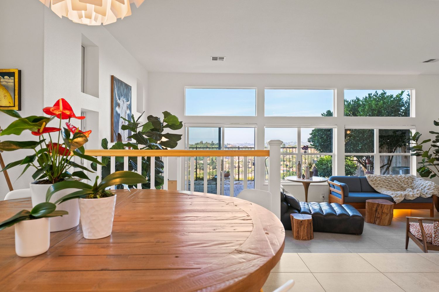 Bright dining area with round wood table, large windows, ocean views, and indoor tropical plants.