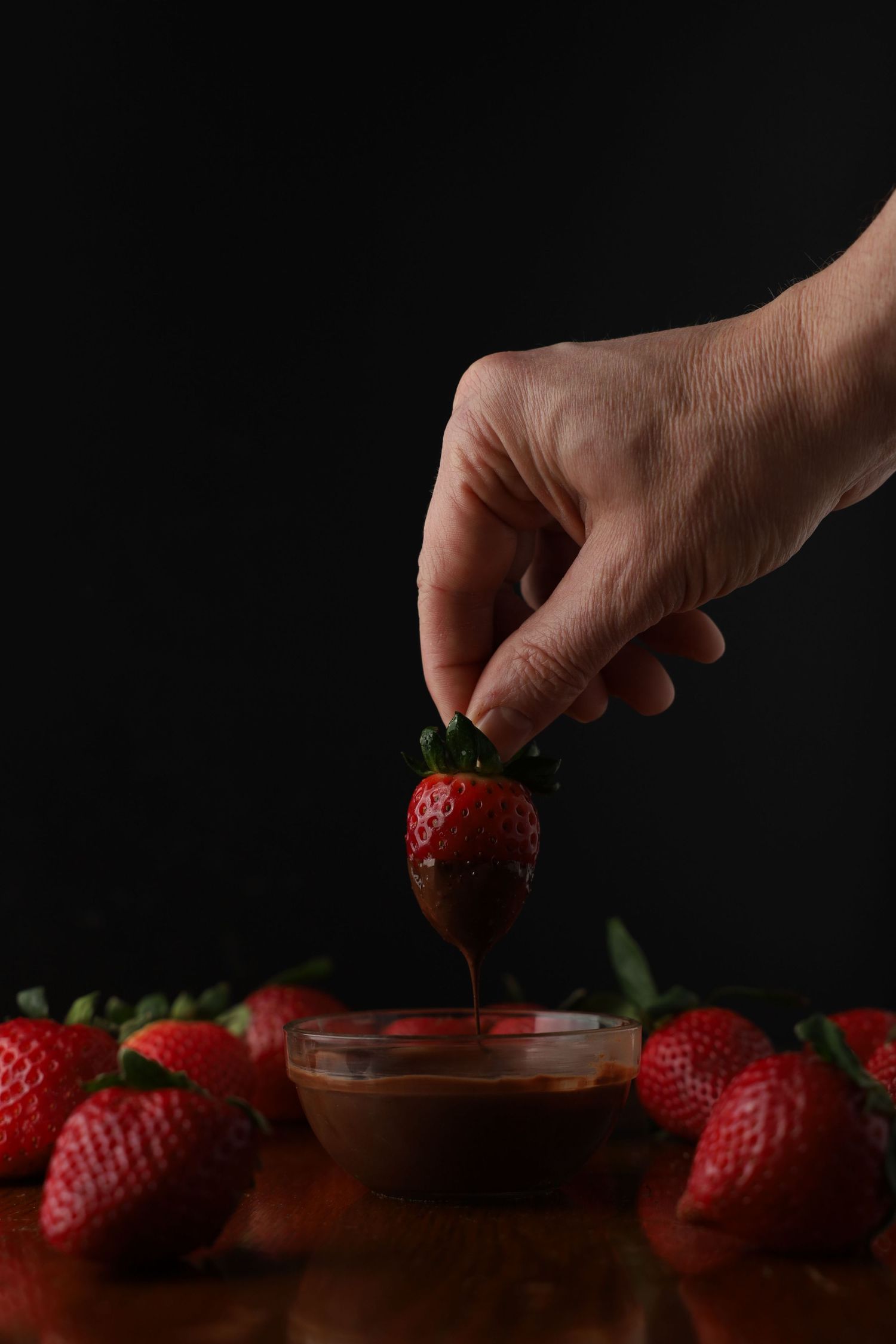 Hand dipping a fresh strawberry into chocolate sauce with strawberries scattered on wooden table.