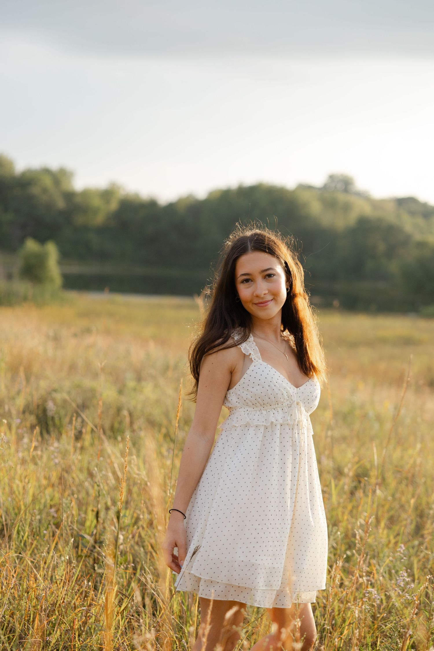 Young woman in white sundress standing in golden field at sunset with trees in background.