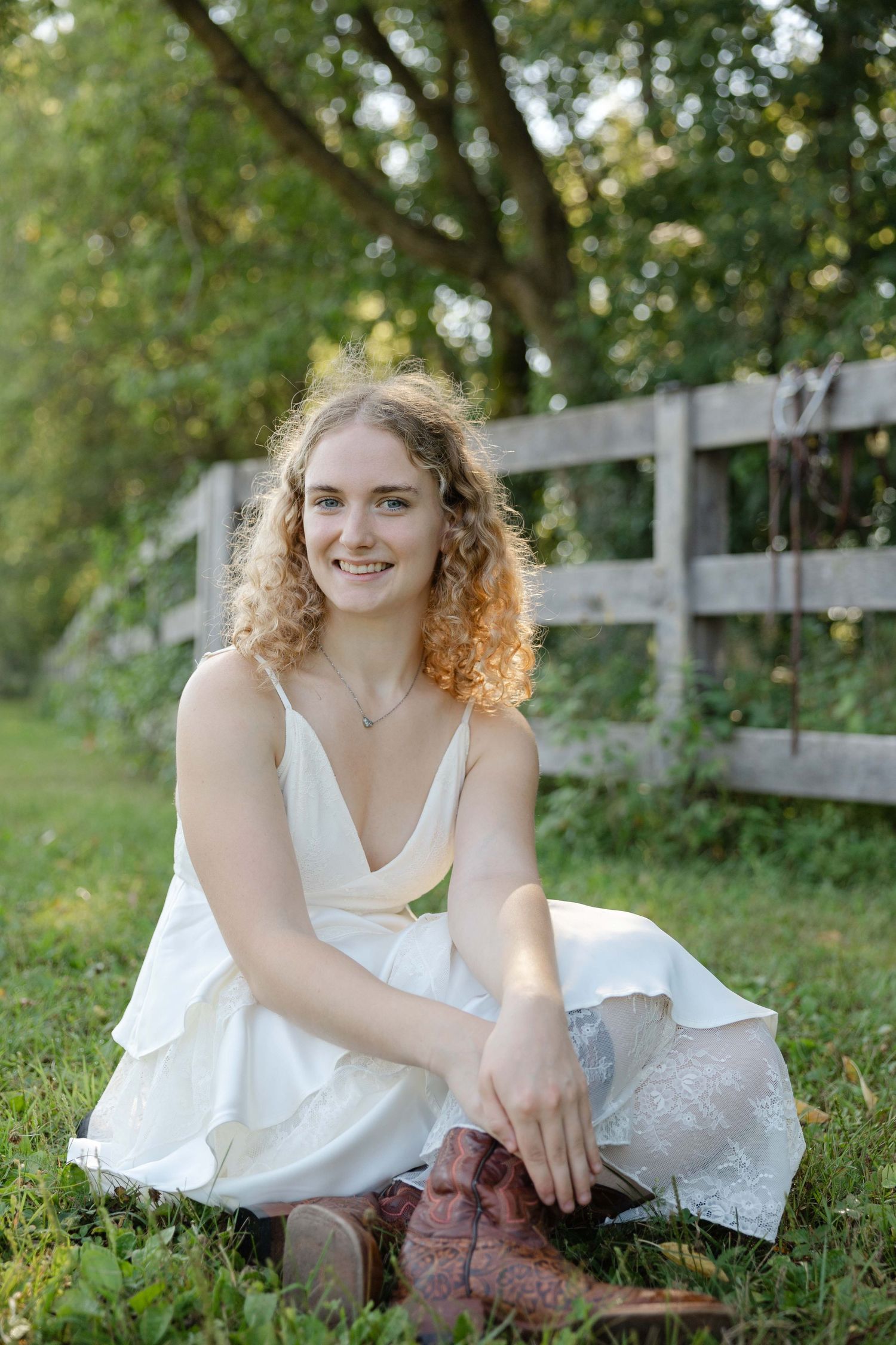 Smiling young woman with curly blonde hair sits on grass in white dress and brown cowboy boots near a wooden fence.