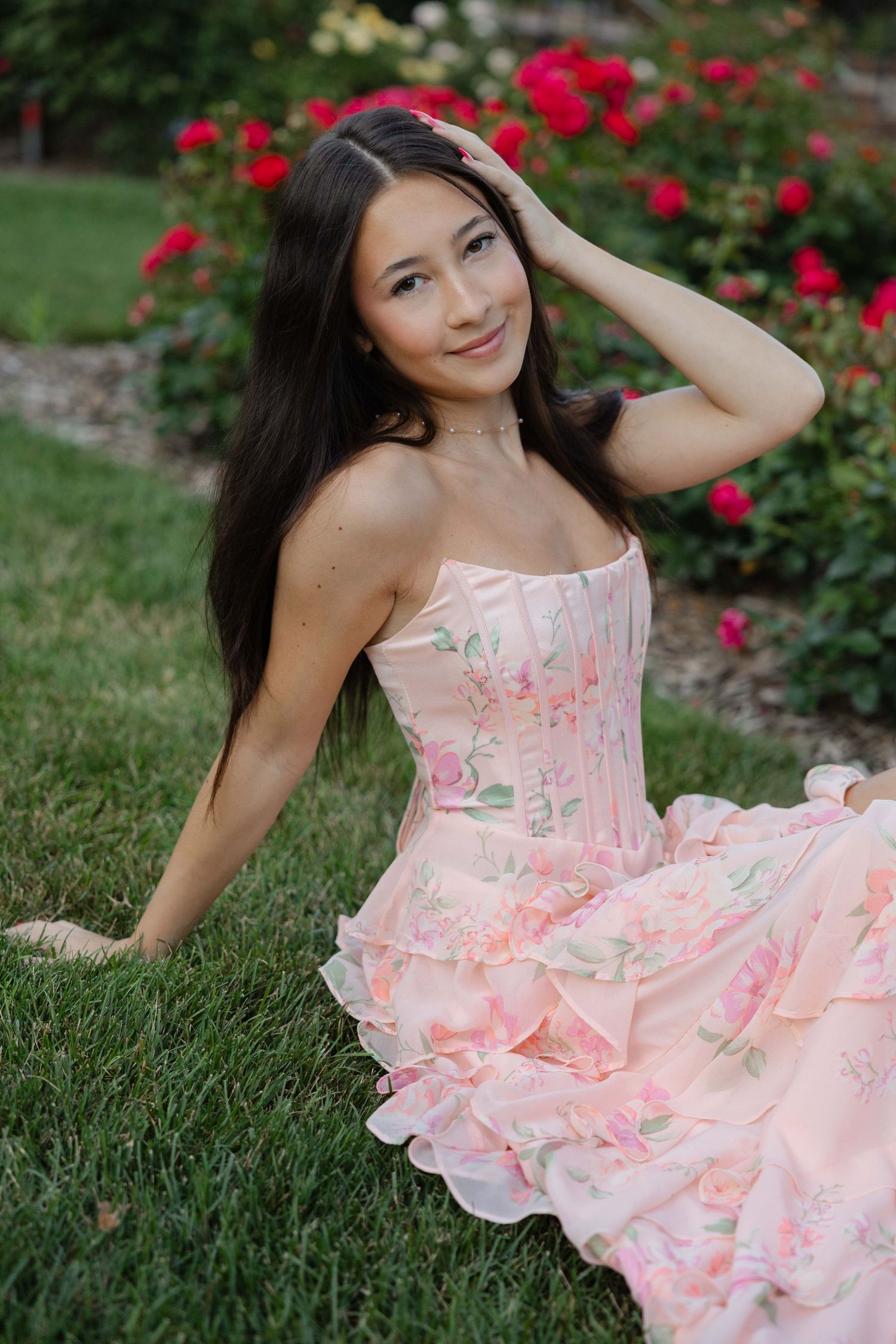 Young woman with long dark hair in pink floral corset gown sits on grass in front of blooming red rose bushes.