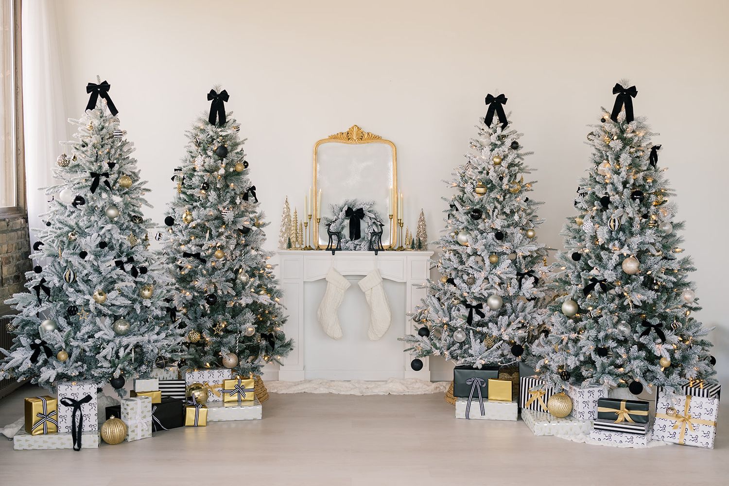 Four flocked Christmas trees with black bows, wrapped gifts and stockings hung on a white fireplace with a gold mirror in a bright room.