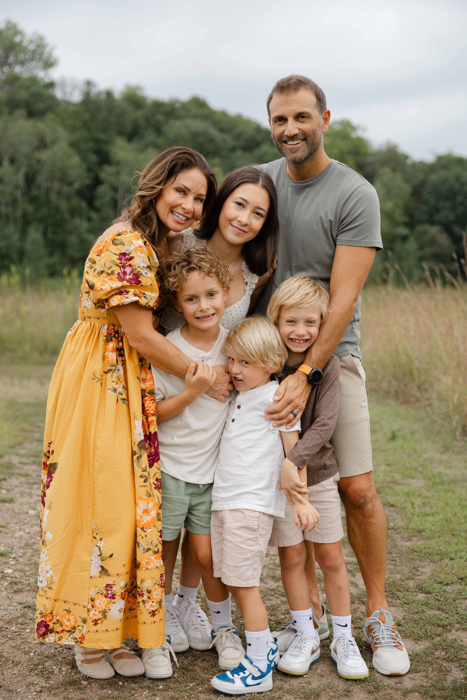 Happy family of five posing outdoors in a field, mom in yellow floral dress, dad in gray tee, three children smiling.