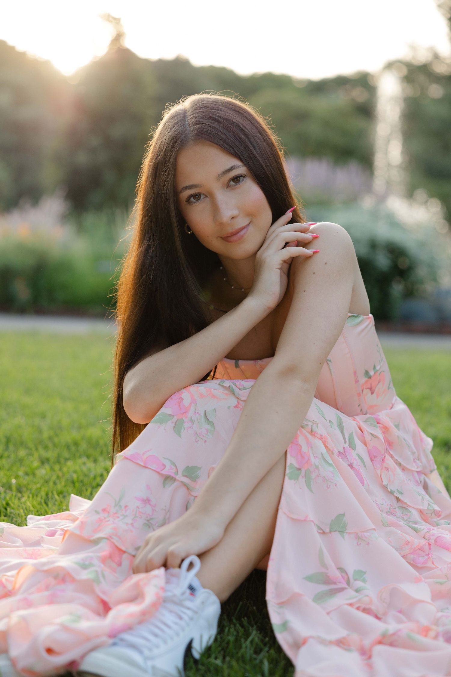 Young woman with long dark hair in pink floral dress sits on grass in a sunlit garden, posing gracefully.
