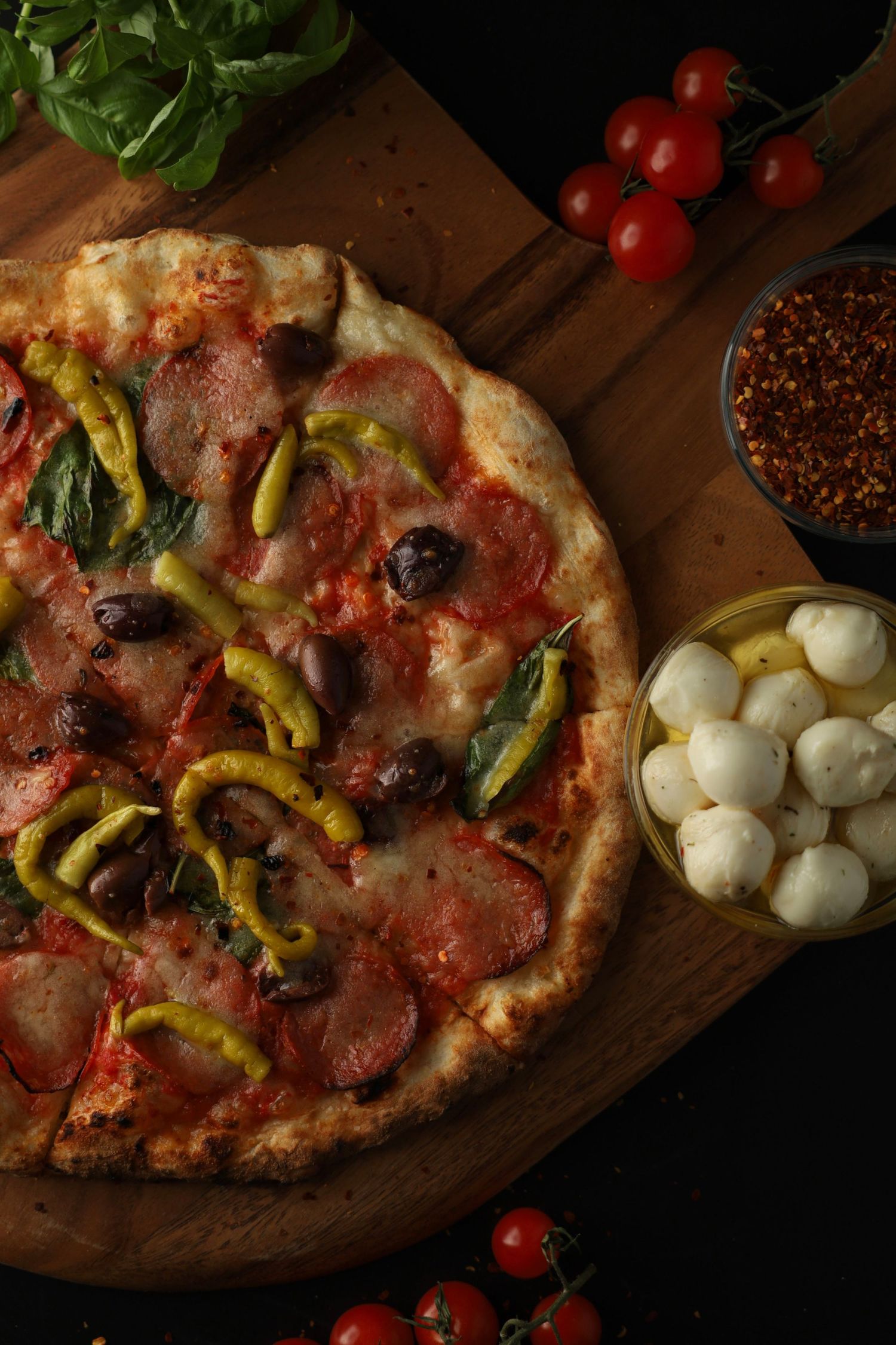 Rustic Italian pizza topped with salami, olives, peppers, and basil served on wooden board with fresh ingredients.