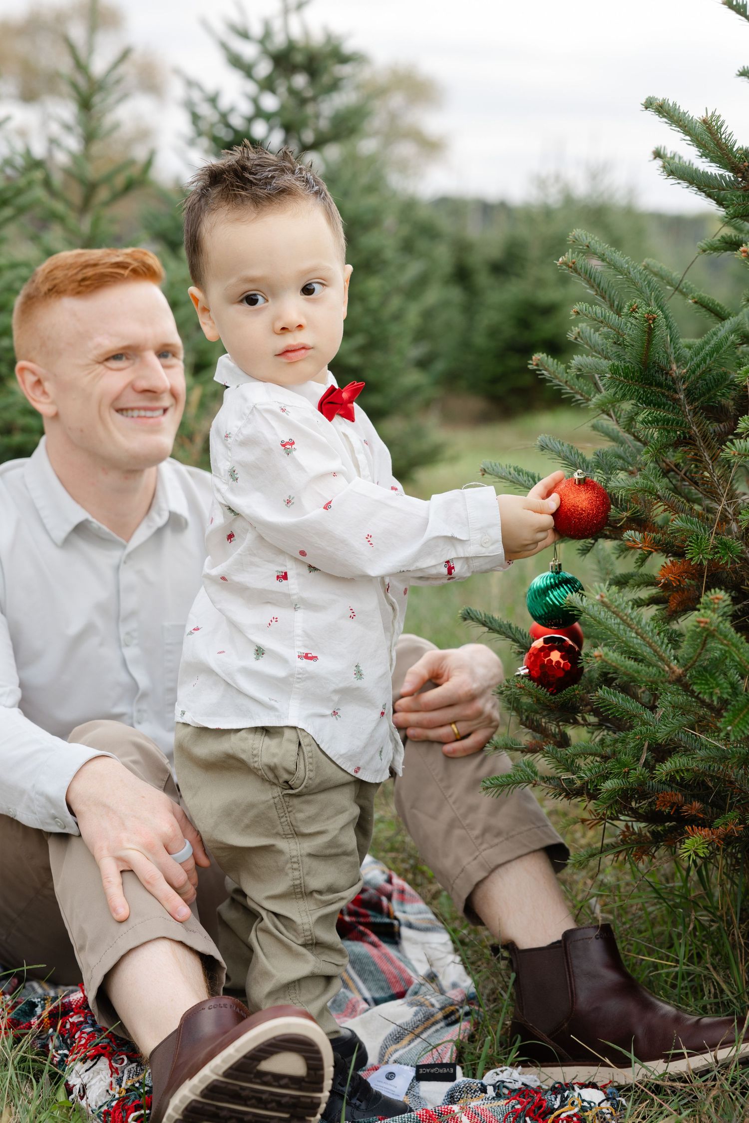 A smiling redhead father sits with his young son decorating a small Christmas tree with red and green ornaments outdoors.