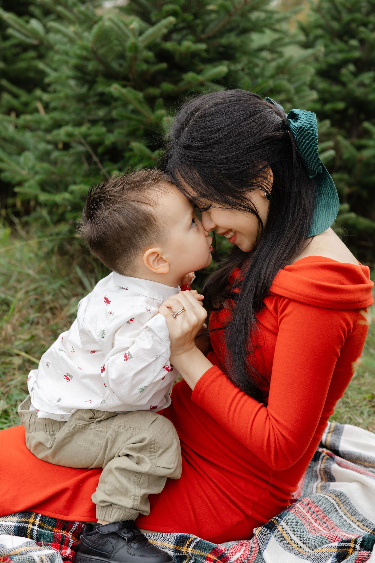 A mother in a red dress tenderly touches noses with her baby boy on a plaid blanket near evergreen trees.