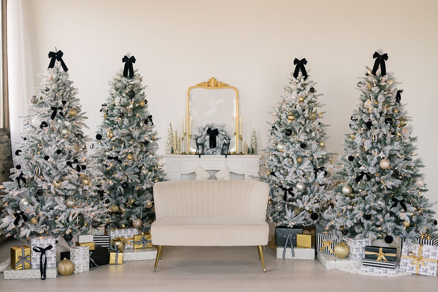Cream sofa between four flocked Christmas trees with black bows and gold ornaments, gift boxes below, and a gold mirror on the wall.