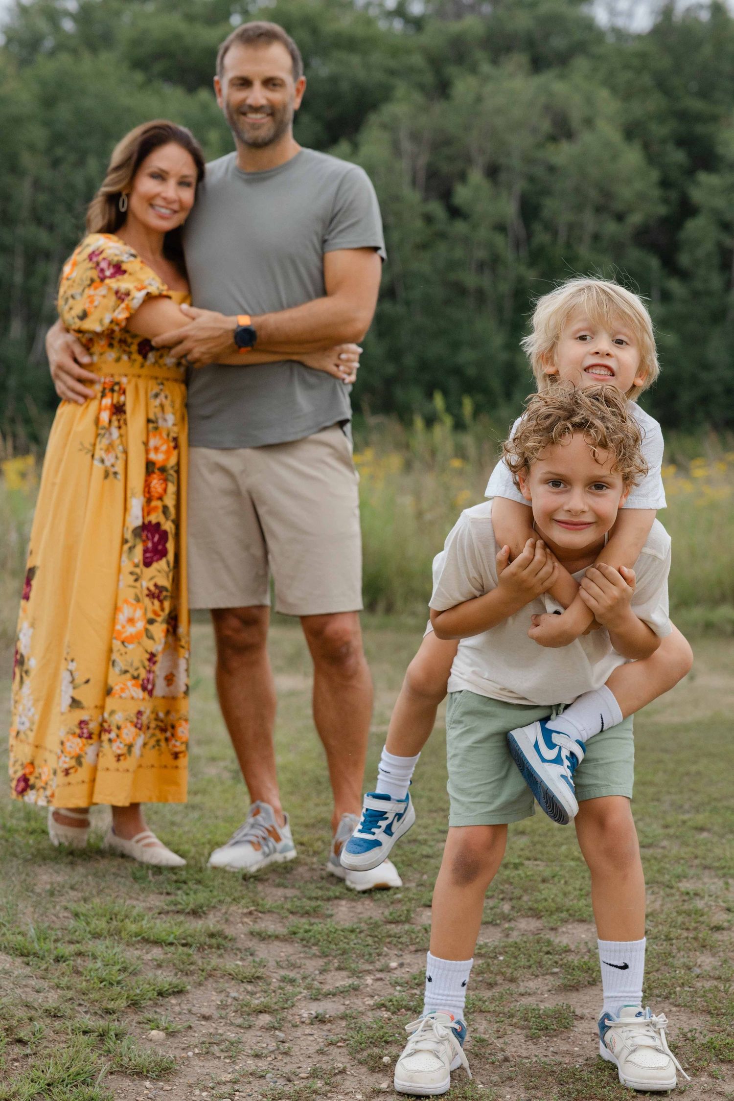 Happy family of four posing outdoors, parents embracing while two young boys give piggyback ride in a green field.