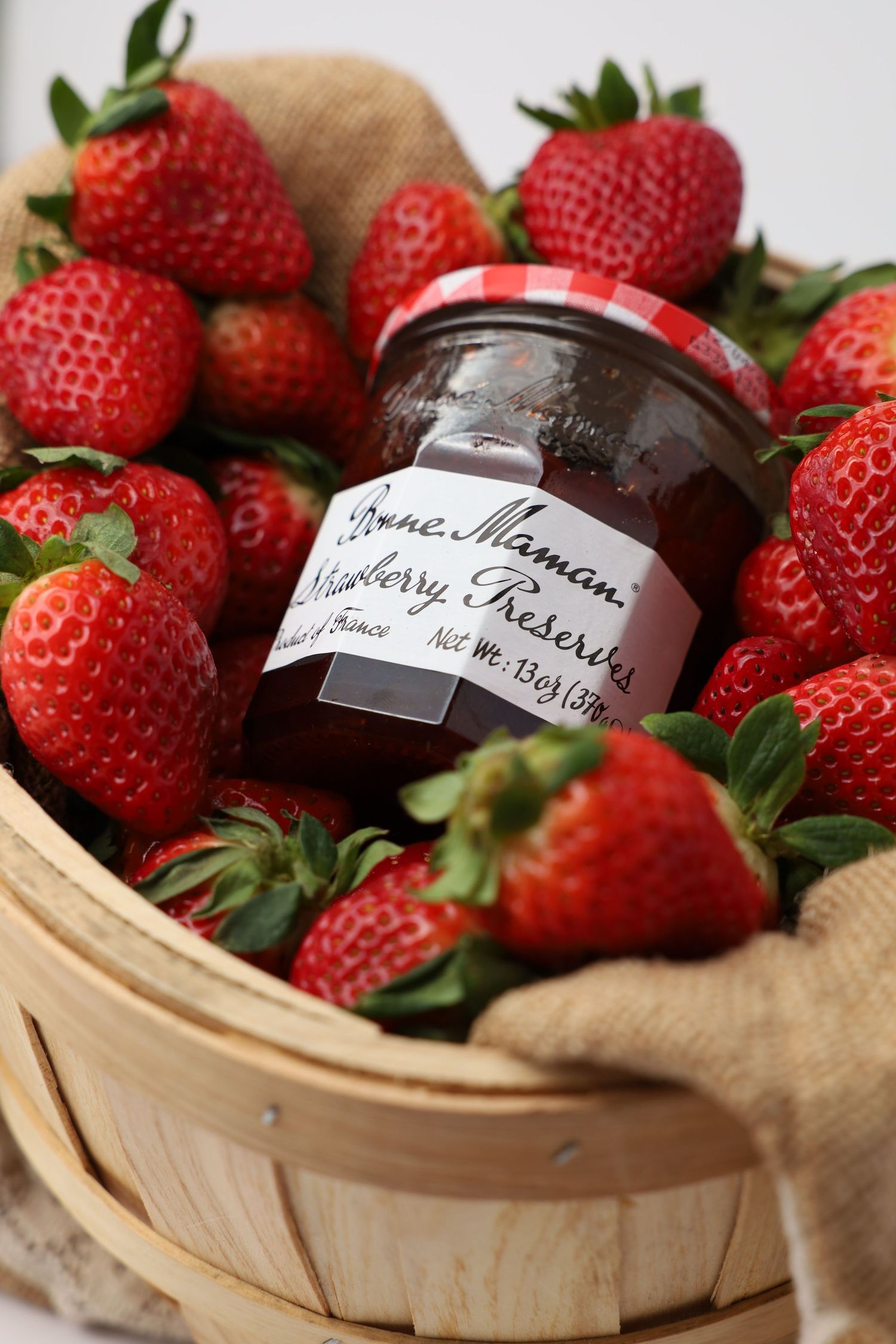 Jar of Bonne Maman strawberry preserves surrounded by fresh ripe strawberries in a wooden basket.