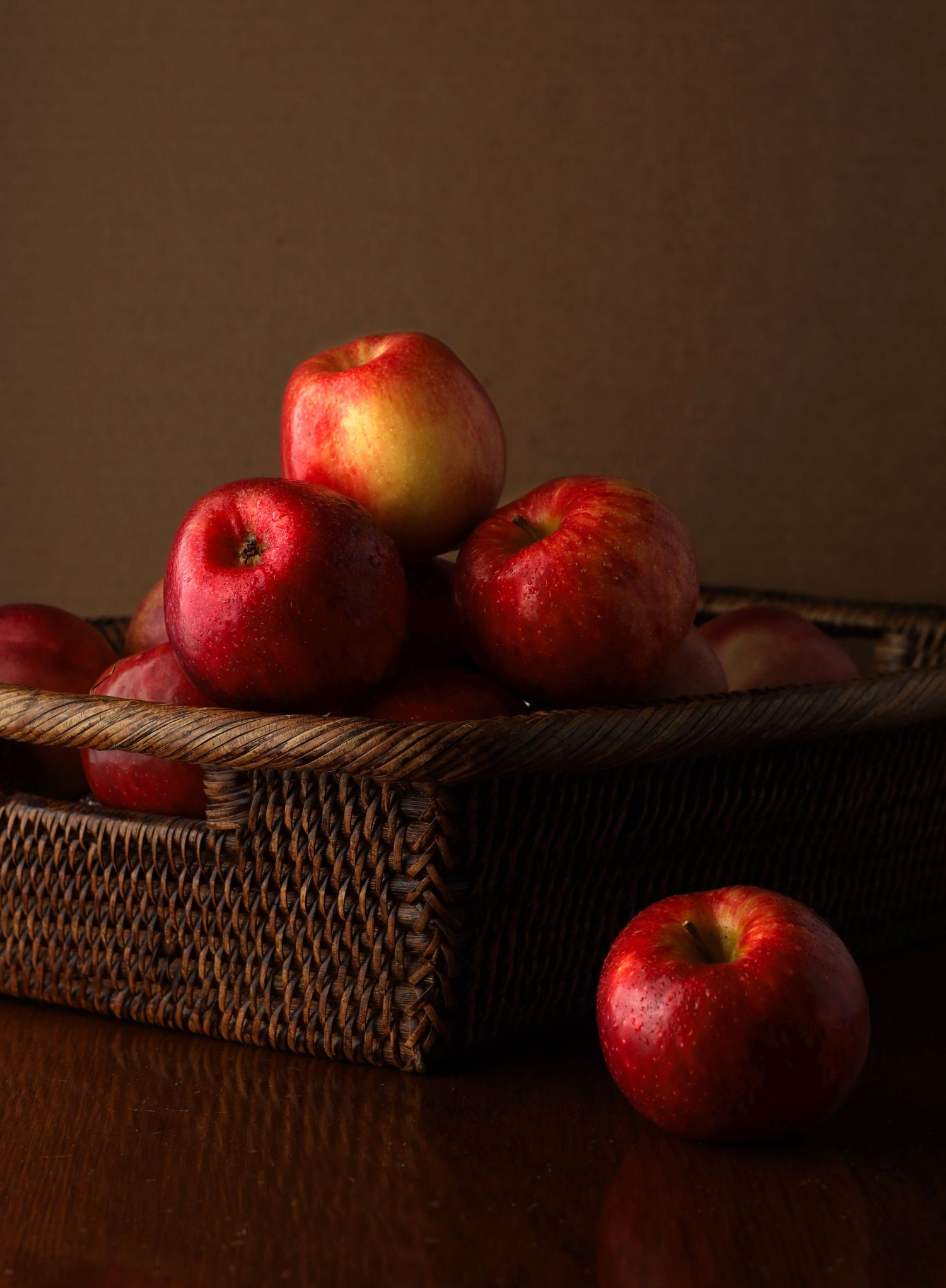 Fresh red apples piled in a dark wicker basket on a wooden table with moody lighting.