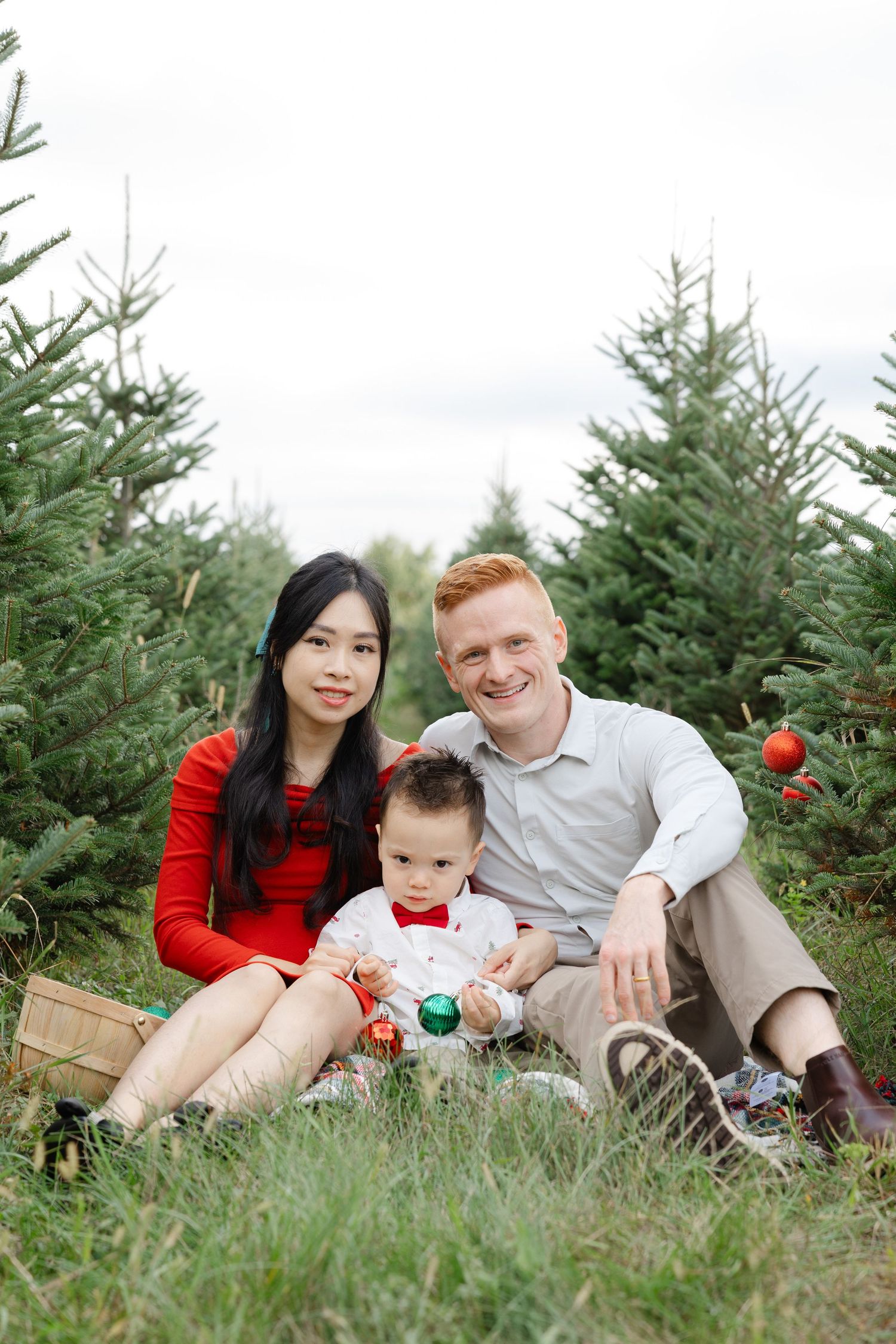 Family of three posing at a Christmas tree farm, with a woman in red, redheaded man, and baby holding ornaments.