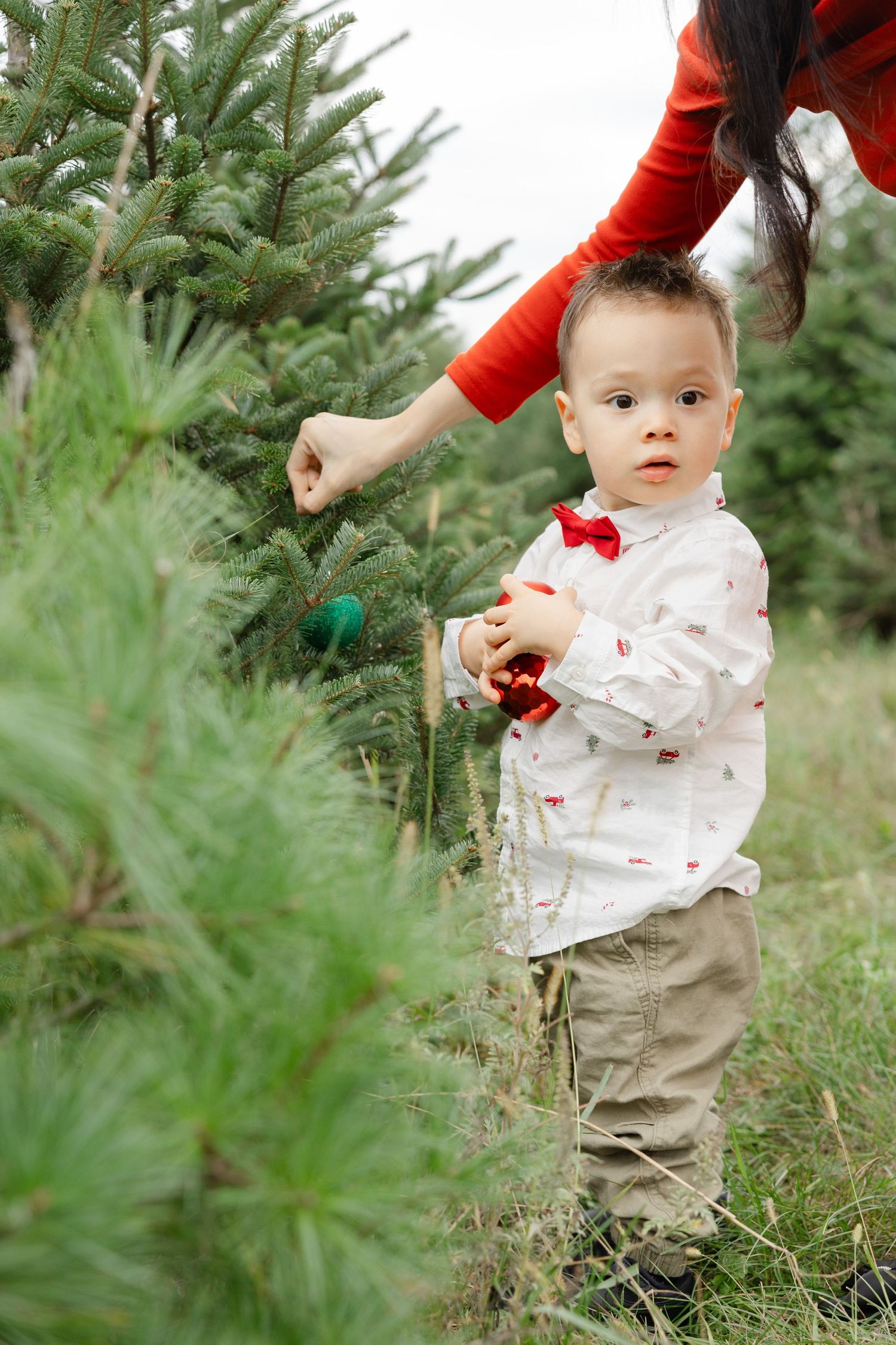 Toddler boy in red bow tie decorating Christmas tree outdoors with mother at a holiday tree farm.