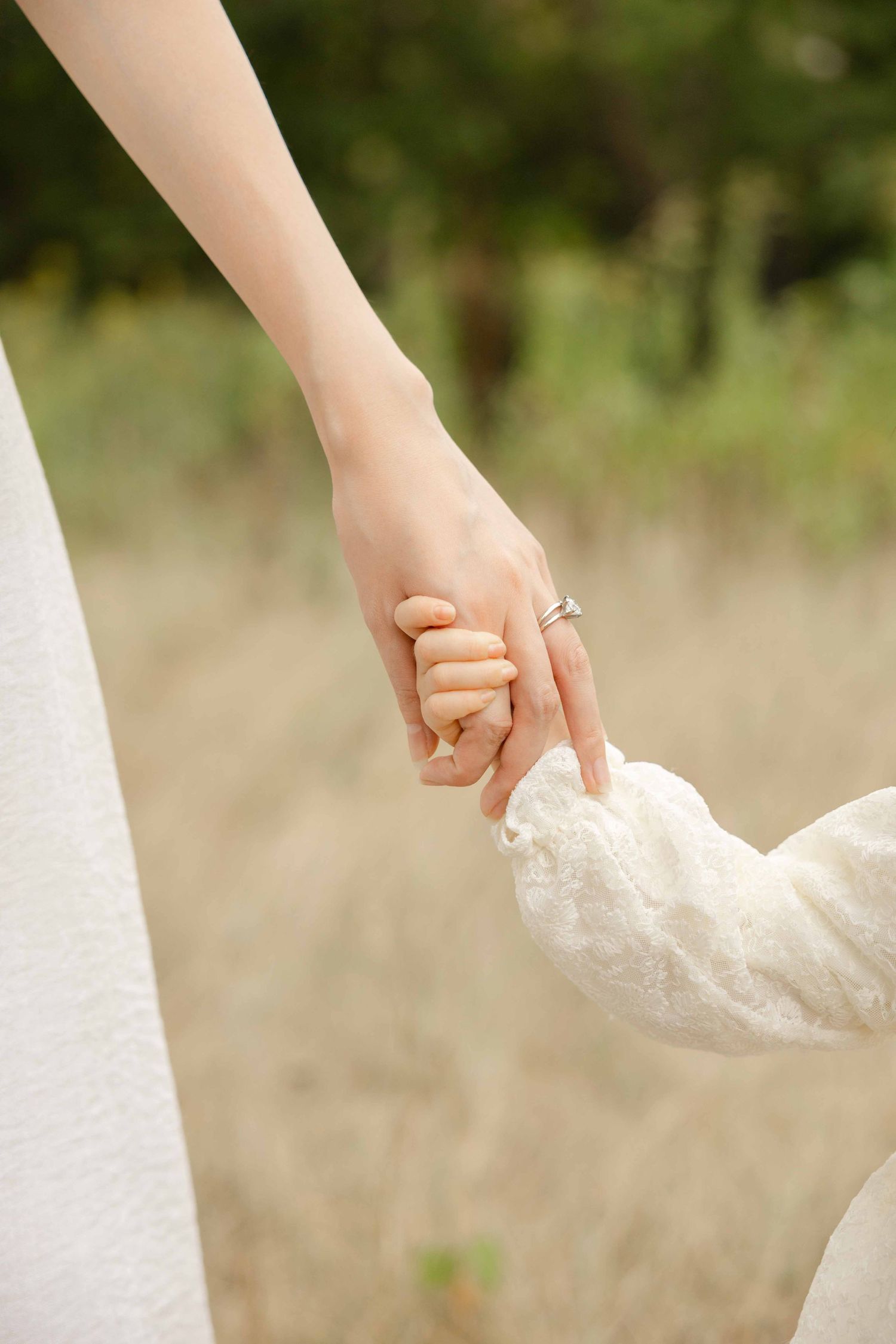 A mother wearing a ring holds her young child's hand outdoors in a natural setting.