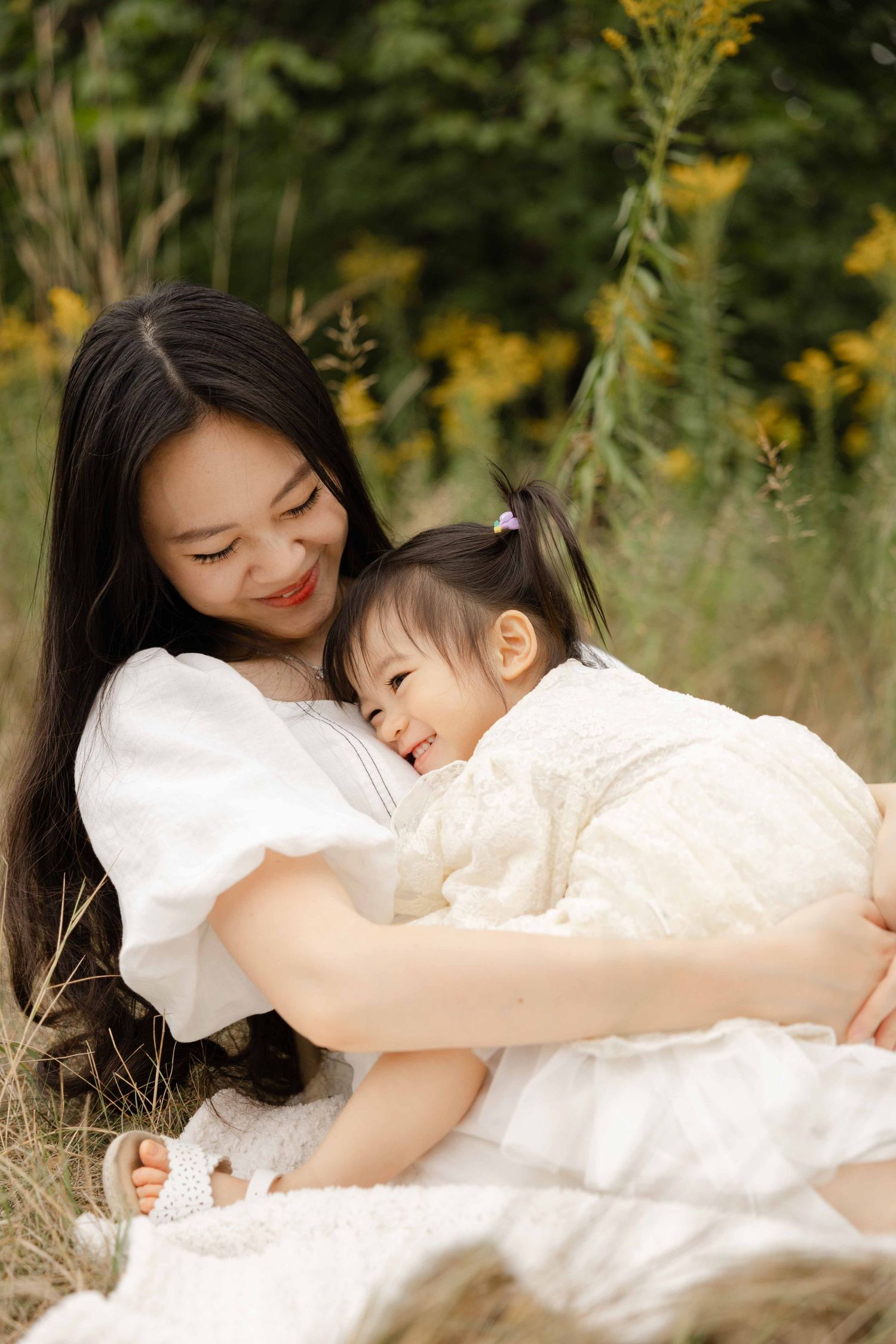 A smiling Asian mother in a white dress cuddles her happy toddler daughter outdoors in a field of yellow flowers.