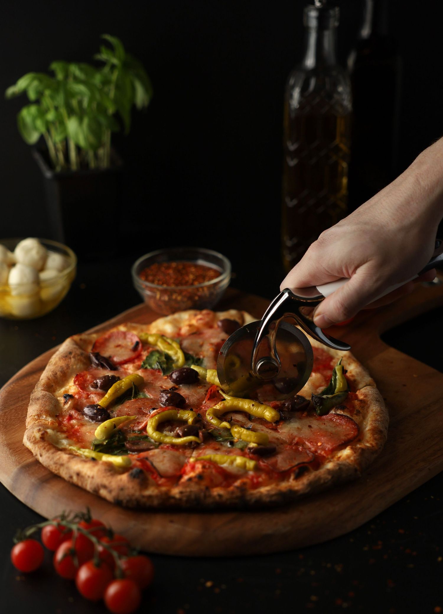 Hand using pizza cutter to slice a rustic Neapolitan pizza with peppers and mushrooms on wooden board.