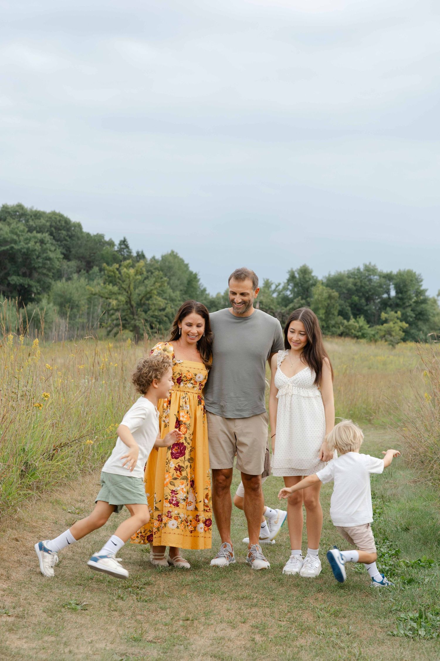 Happy family of five enjoying a sunny day outdoors in a meadow, parents smiling as children playfully run around them.
