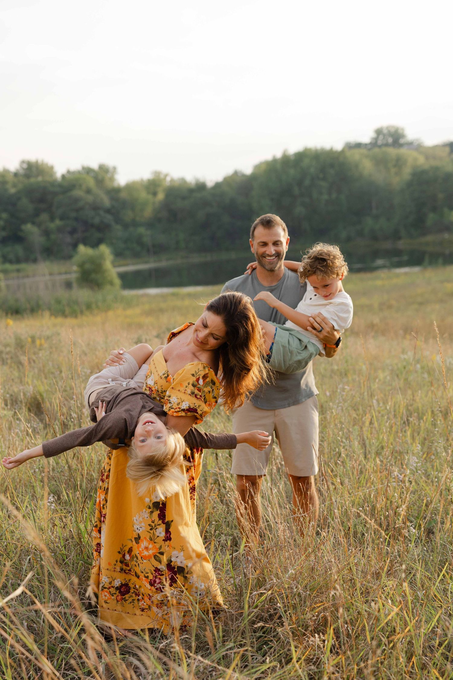 Happy family playing in a sunny meadow, mother swinging child while father holds toddler, with trees and lake in background.