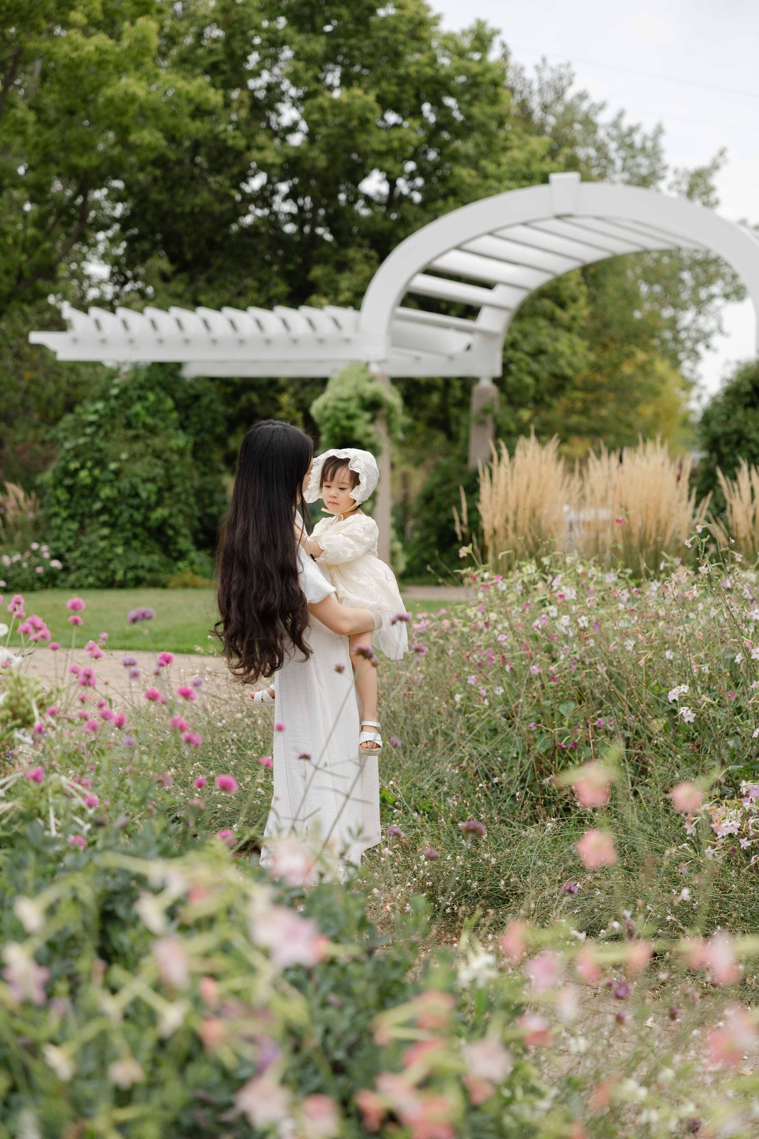 A mother in a white dress holds her baby girl in a blooming garden with a white pergola arch in the background.