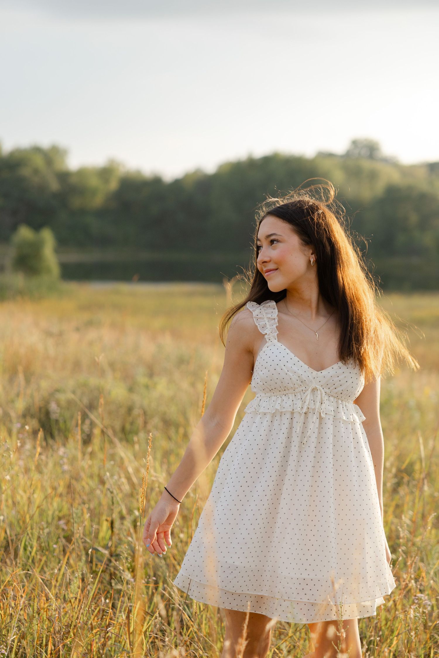 Young woman in white sundress walking through golden field at sunset with trees and lake in background.