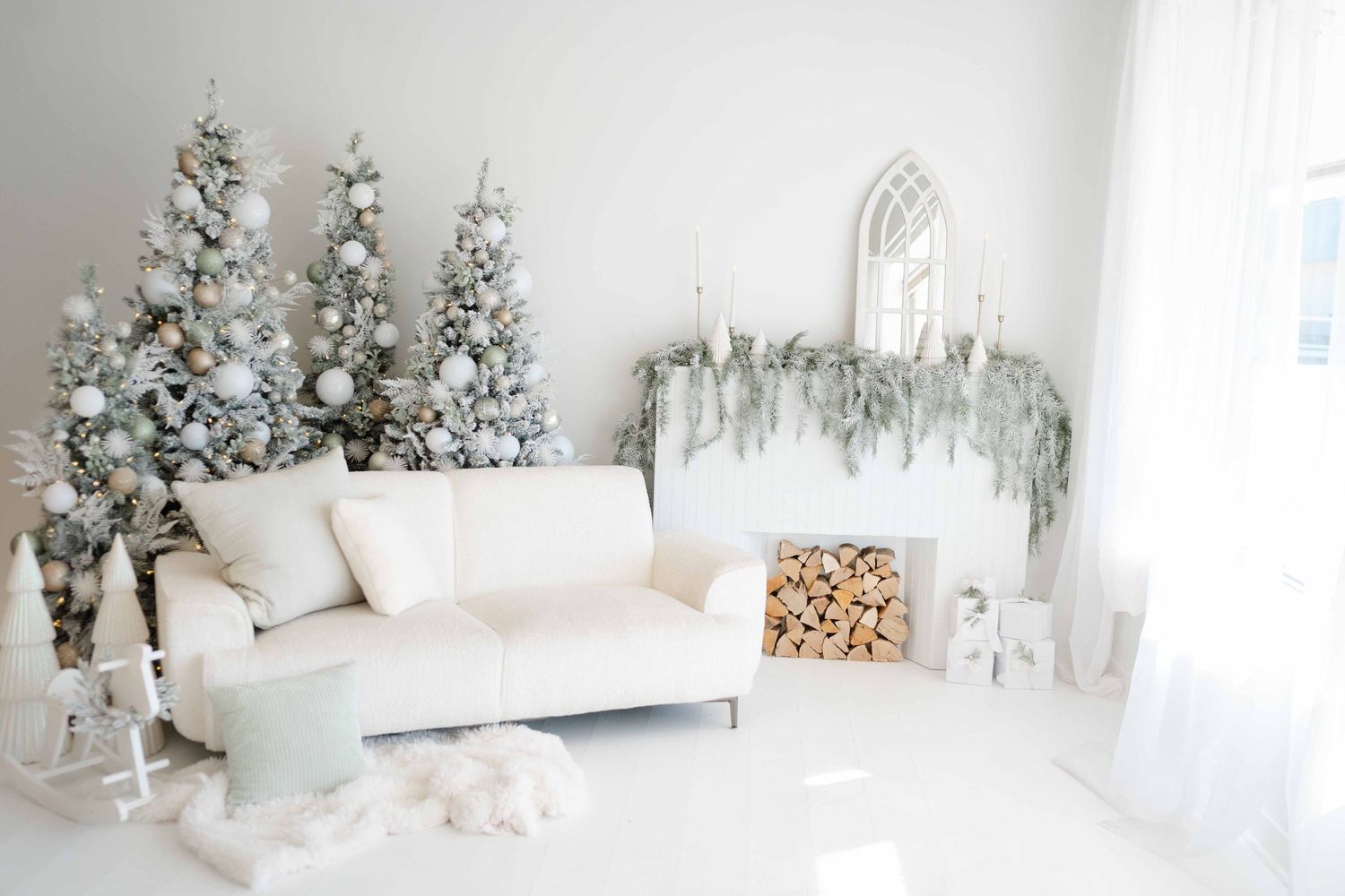 White living room with snow-flocked Christmas trees, white sofa, fireplace mantel with greenery, and stacked firewood decor.
