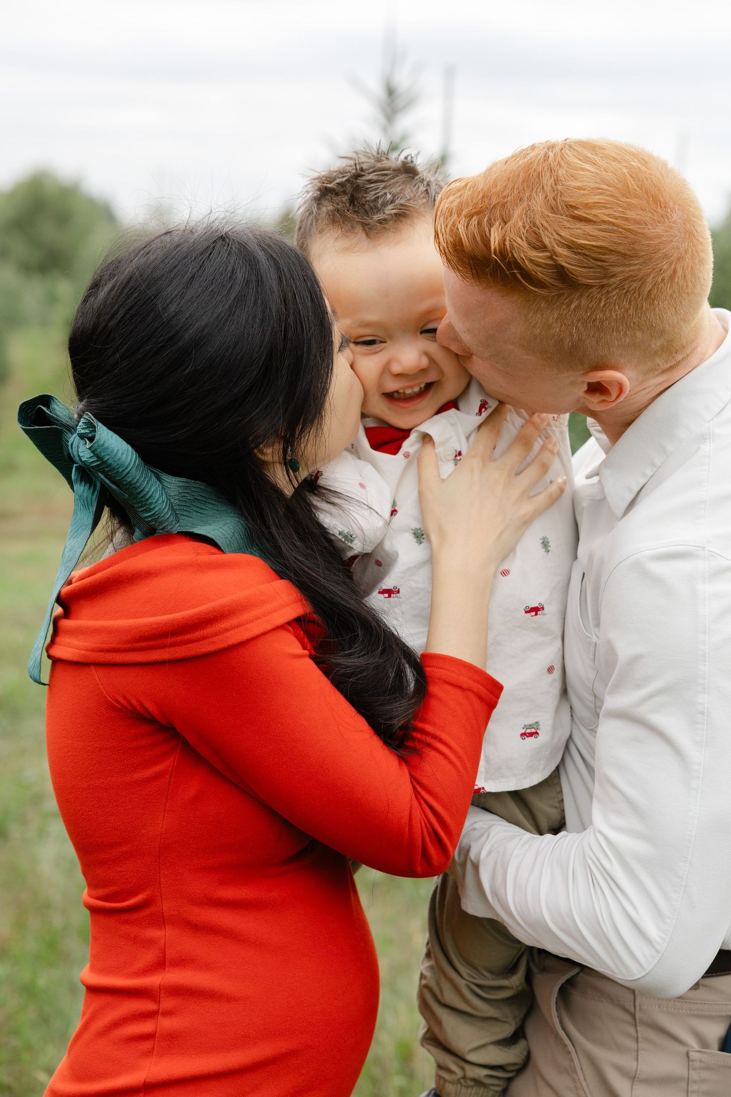 A happy family with a redhead father and brunette mother in a red dress kissing their laughing toddler son outdoors.