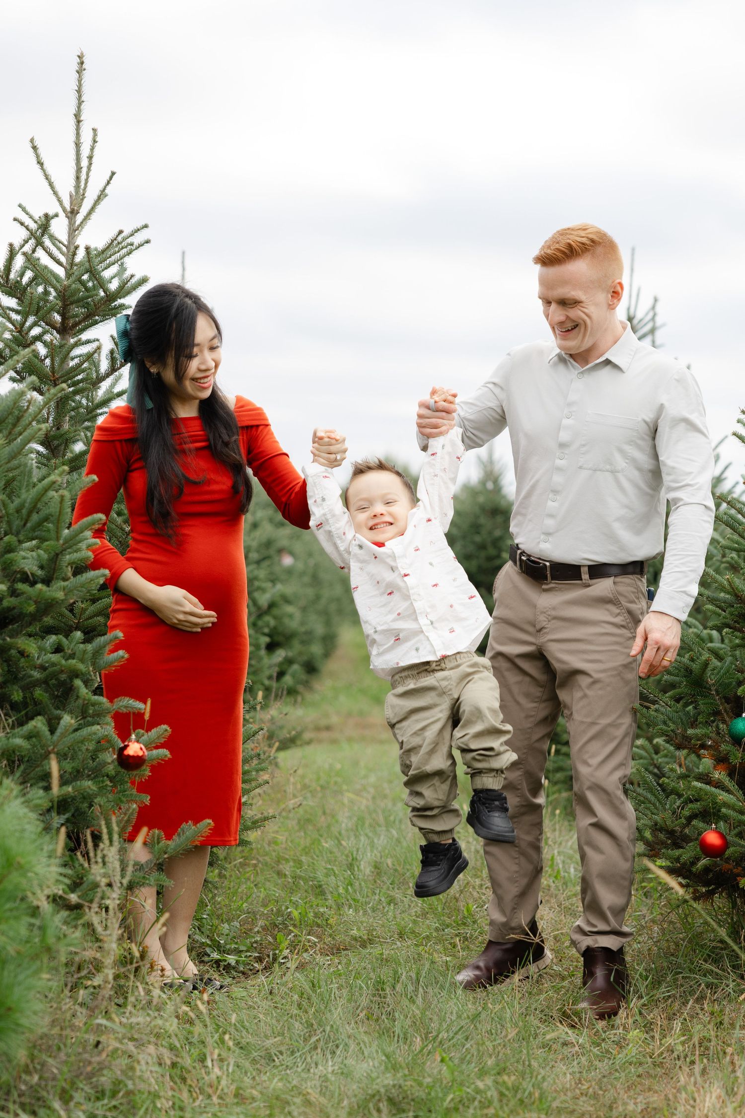 A happy family swings their toddler between them at a Christmas tree farm, mom in red dress, dad in gray shirt.