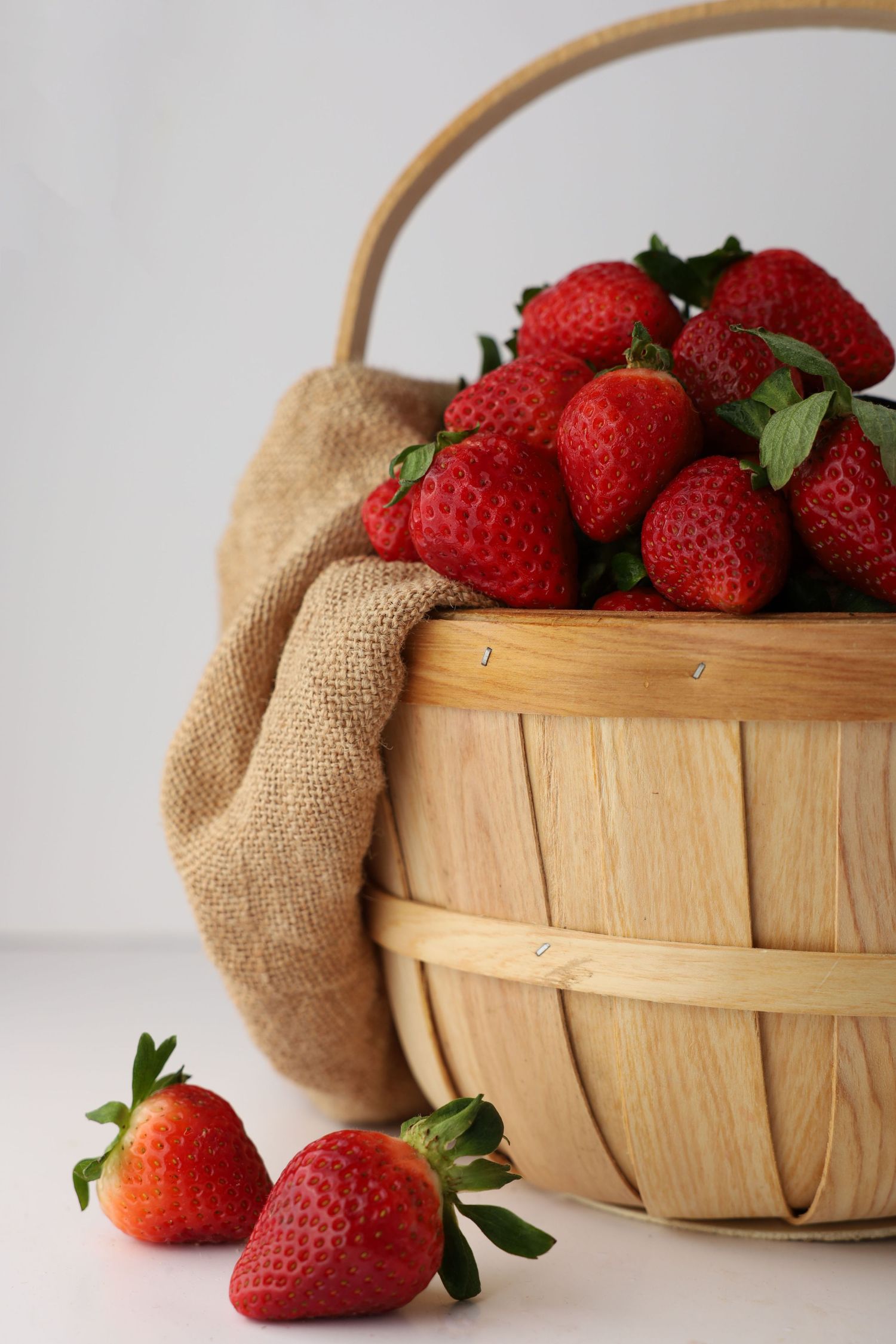 Fresh red strawberries overflowing from a wooden basket with burlap lining on white surface.
