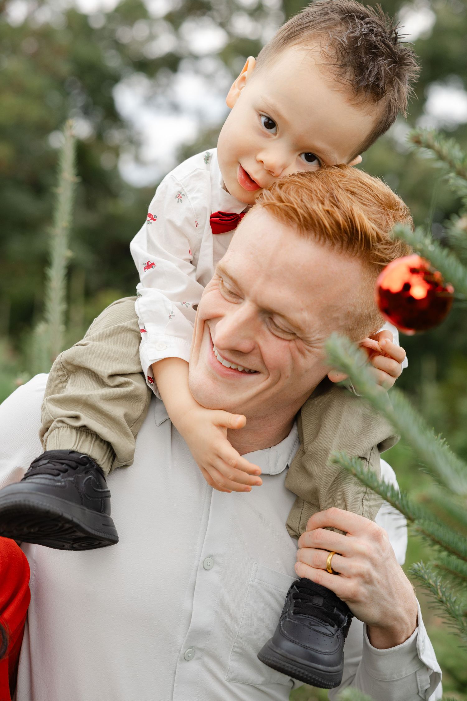 A smiling redhead father carries his young son on his shoulders near a decorated Christmas tree outdoors.