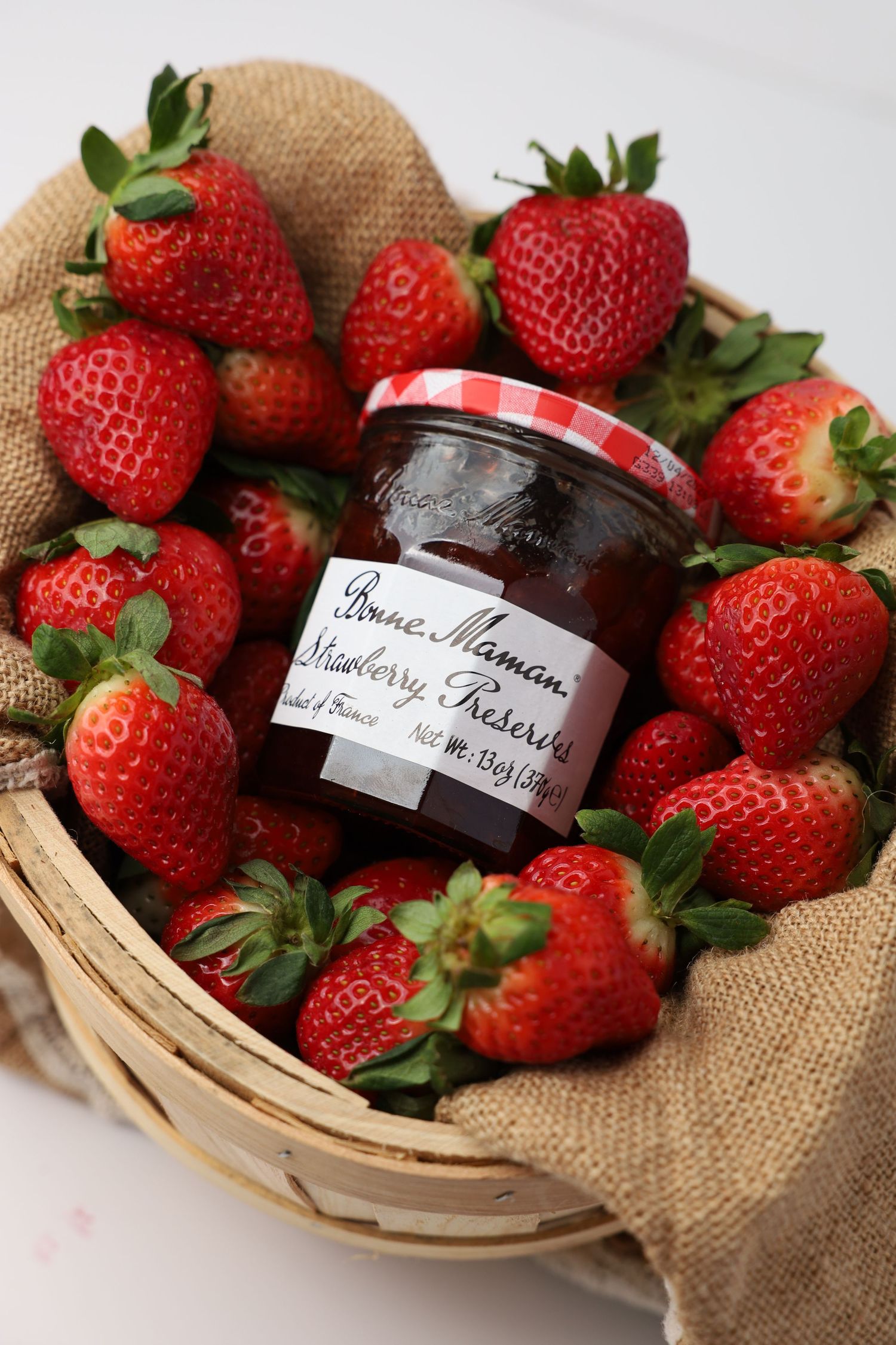 Jar of Bonne Maman strawberry preserves nestled among fresh red strawberries in a burlap-lined basket.