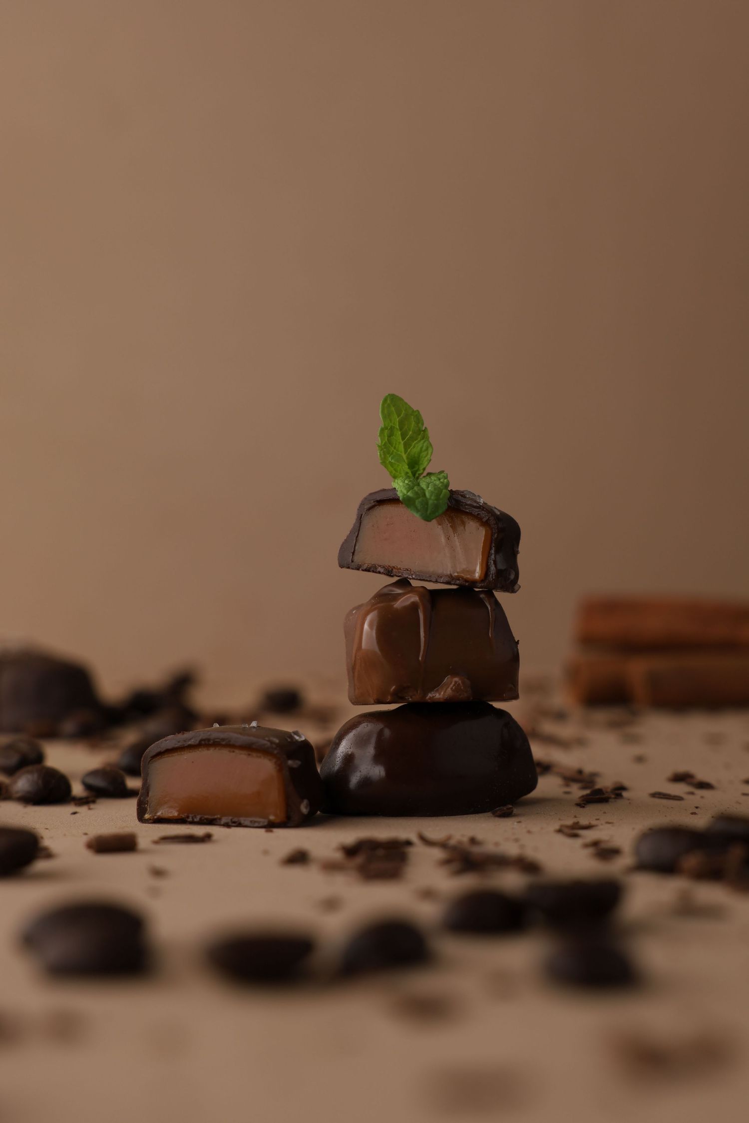 Stack of three chocolate candies topped with mint leaves surrounded by coffee beans on beige background.