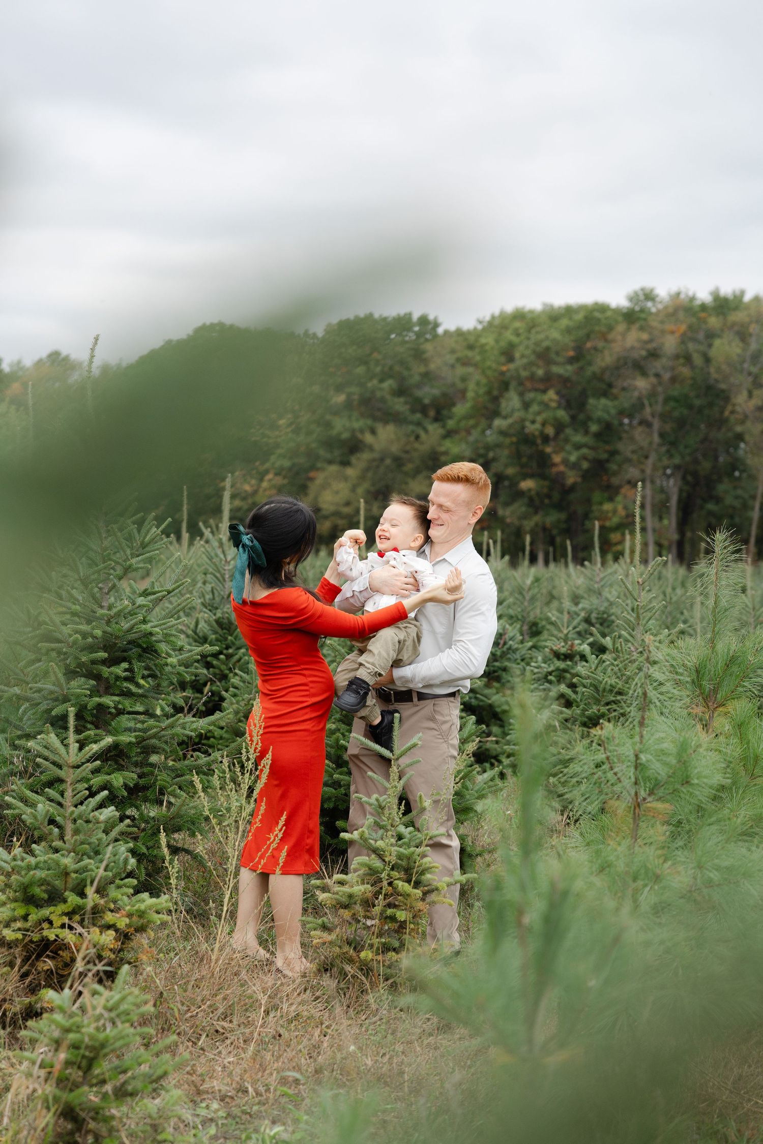 A joyful family with a baby at a Christmas tree farm, woman in red dress and man with red hair outdoors.