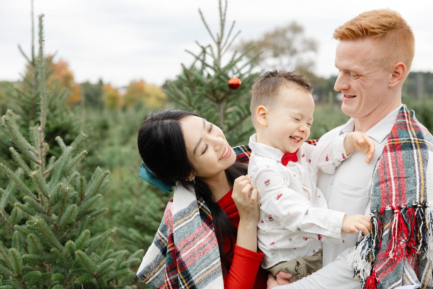 Family smiling together at a Christmas tree farm, wrapped in plaid blankets, holding a young child in a red bow tie.