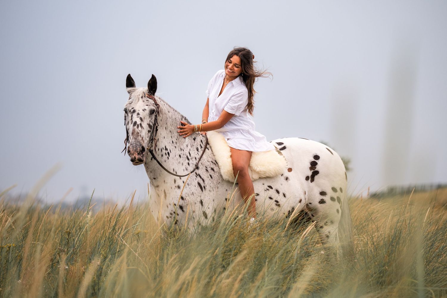 Young woman in white dress riding a spotted Appaloosa horse through a golden grass field under a cloudy sky.