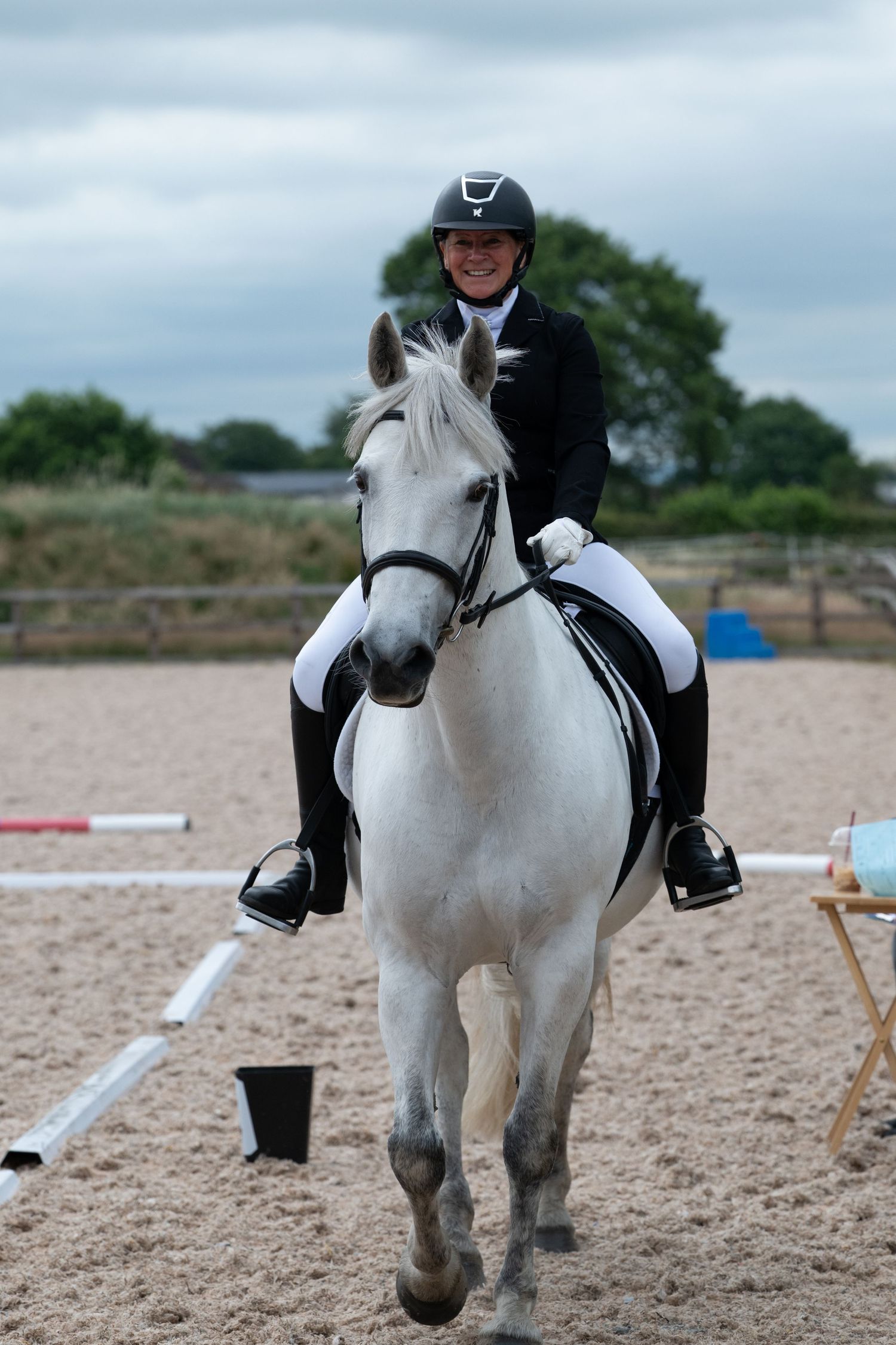 Smiling equestrian in black jacket and helmet rides white horse in sandy arena during show jumping competition.