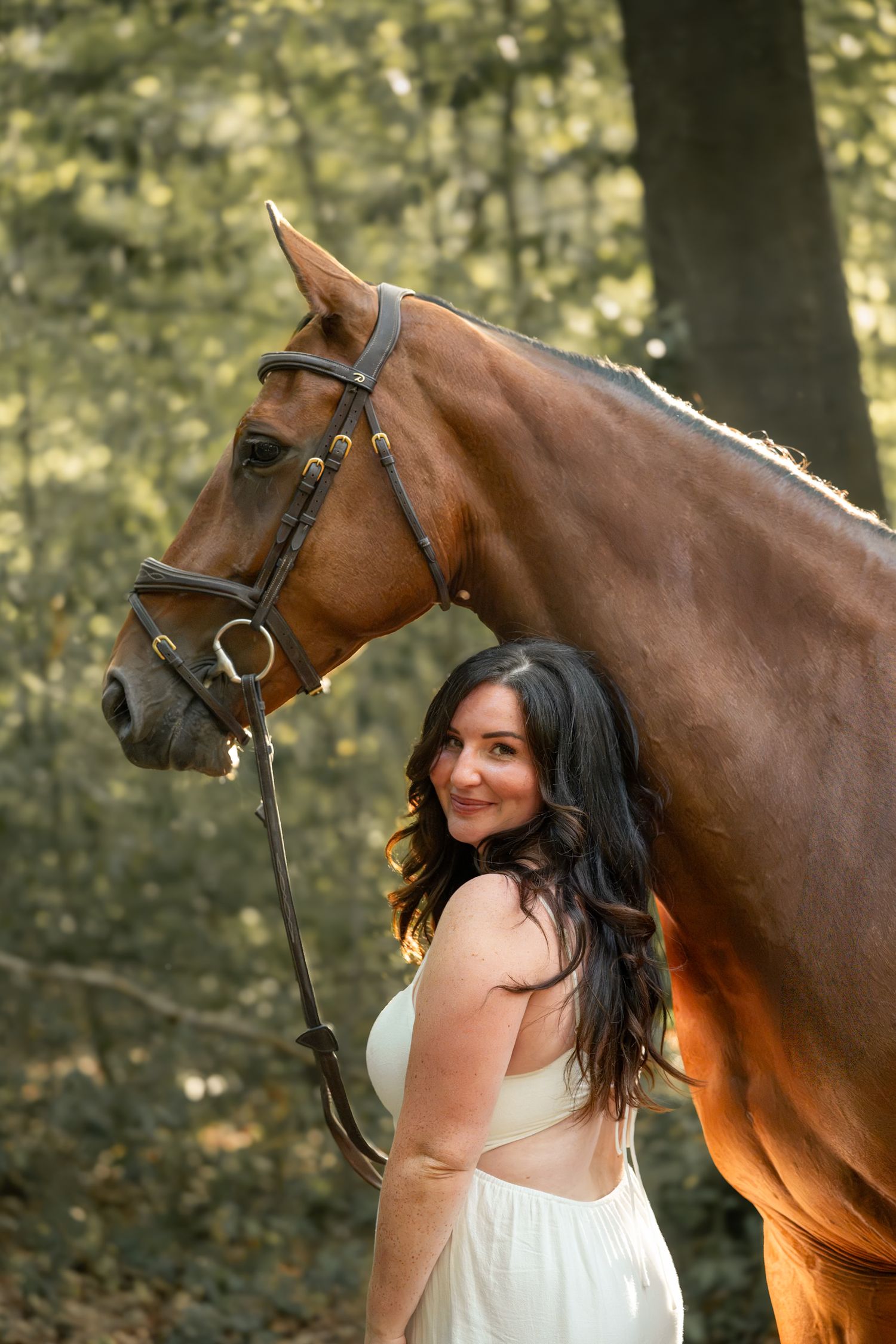 A young woman in a white dress smiles beside a chestnut horse with a bridle in a sunlit forest.