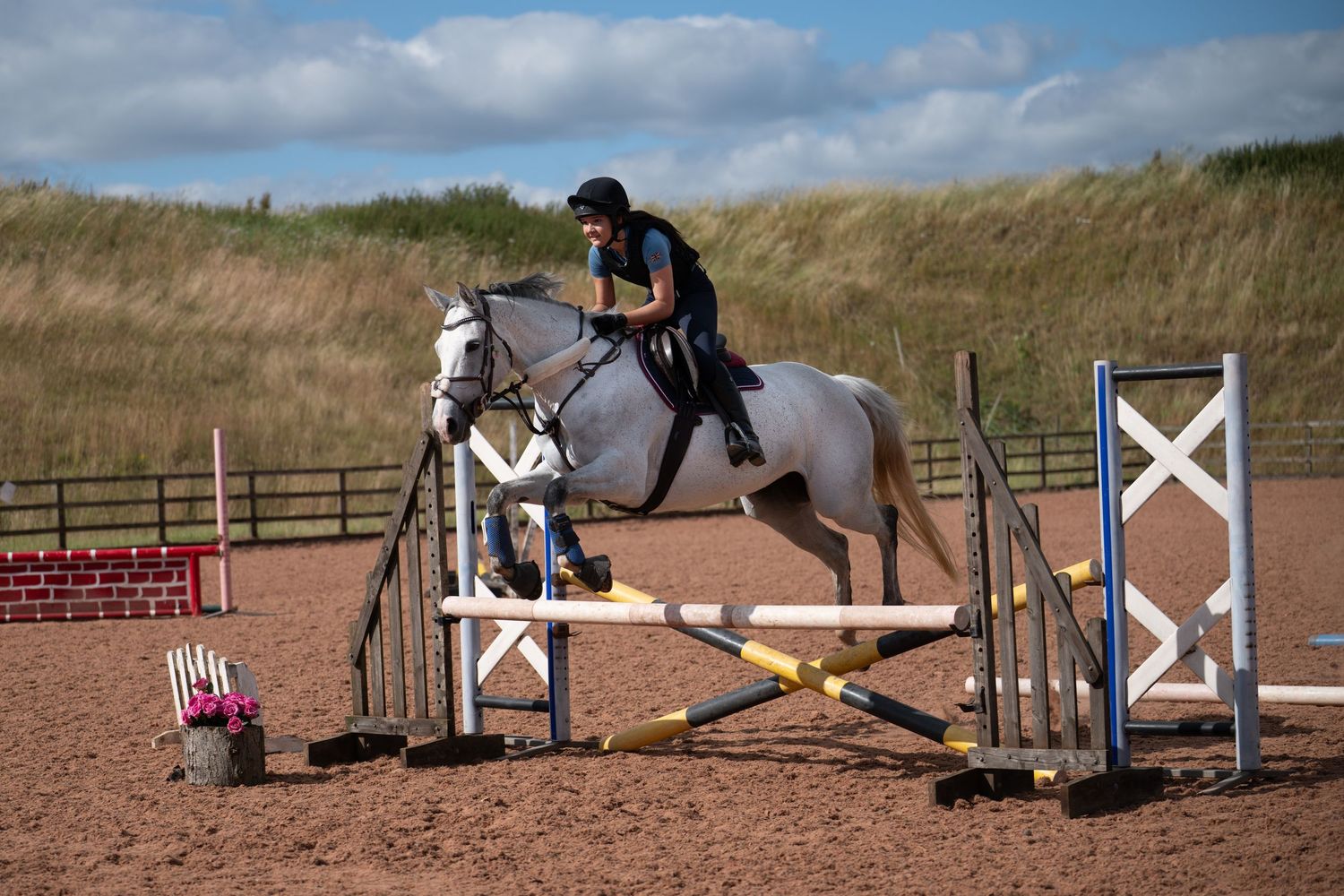 An equestrian in black rides a grey horse over a jump in a sandy outdoor arena under blue skies.