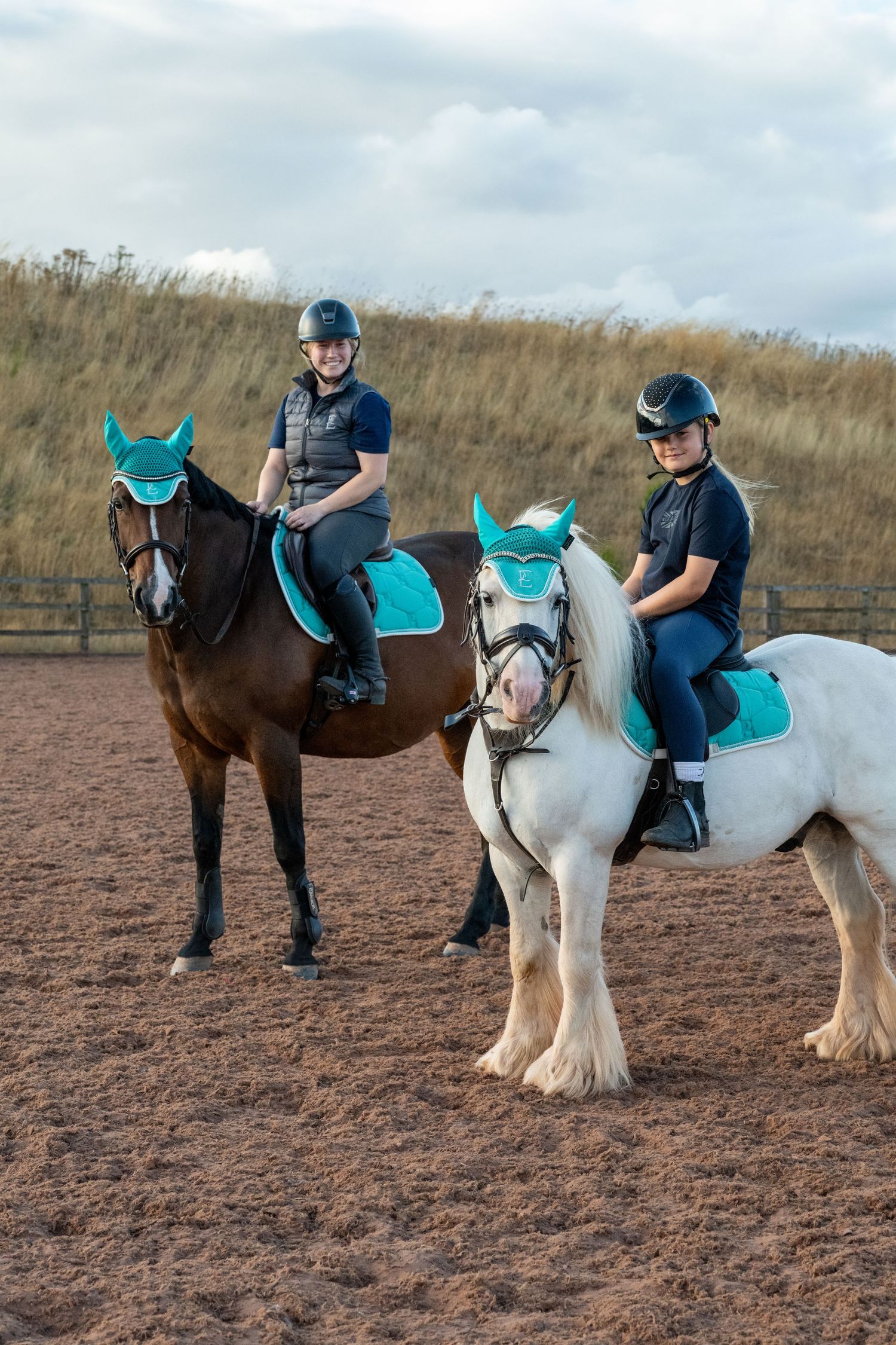 Two young riders in helmets sit on brown and white horses with teal saddle pads in an outdoor riding arena.